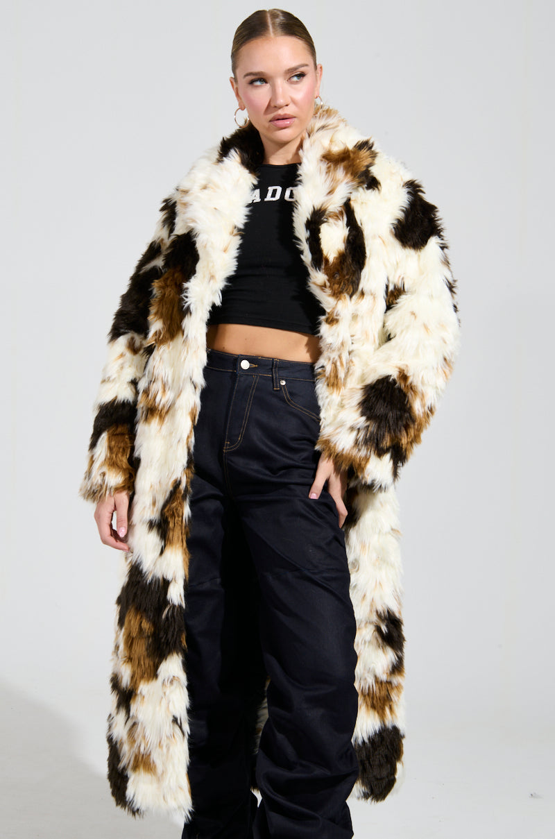 Dressed in the BRONX FAUX FUR COAT, with its black and white design, an individual stands confidently. Beneath the coat is a black crop top paired with dark pants. The plain background accentuates their shaggy-chic style.