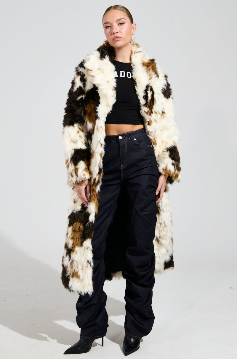 Dressed in the stylish BRONX FAUX FUR COAT in a long calico colorway, an individual exudes confidence. They pair it with a black crop top and dark pants, finished off with pointed black boots. A plain background highlights their shaggy-chic, fashion-forward ensemble.