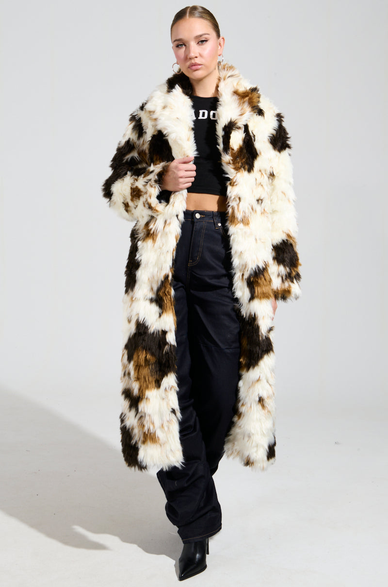 A person stands confidently wearing the BRONX FAUX FUR COAT, showcasing a long, shaggy-chic design in a calico colorway of white, brown, and black. They pair this stylish coat with a black crop top and pants, complemented by matching boots against a plain gray background.