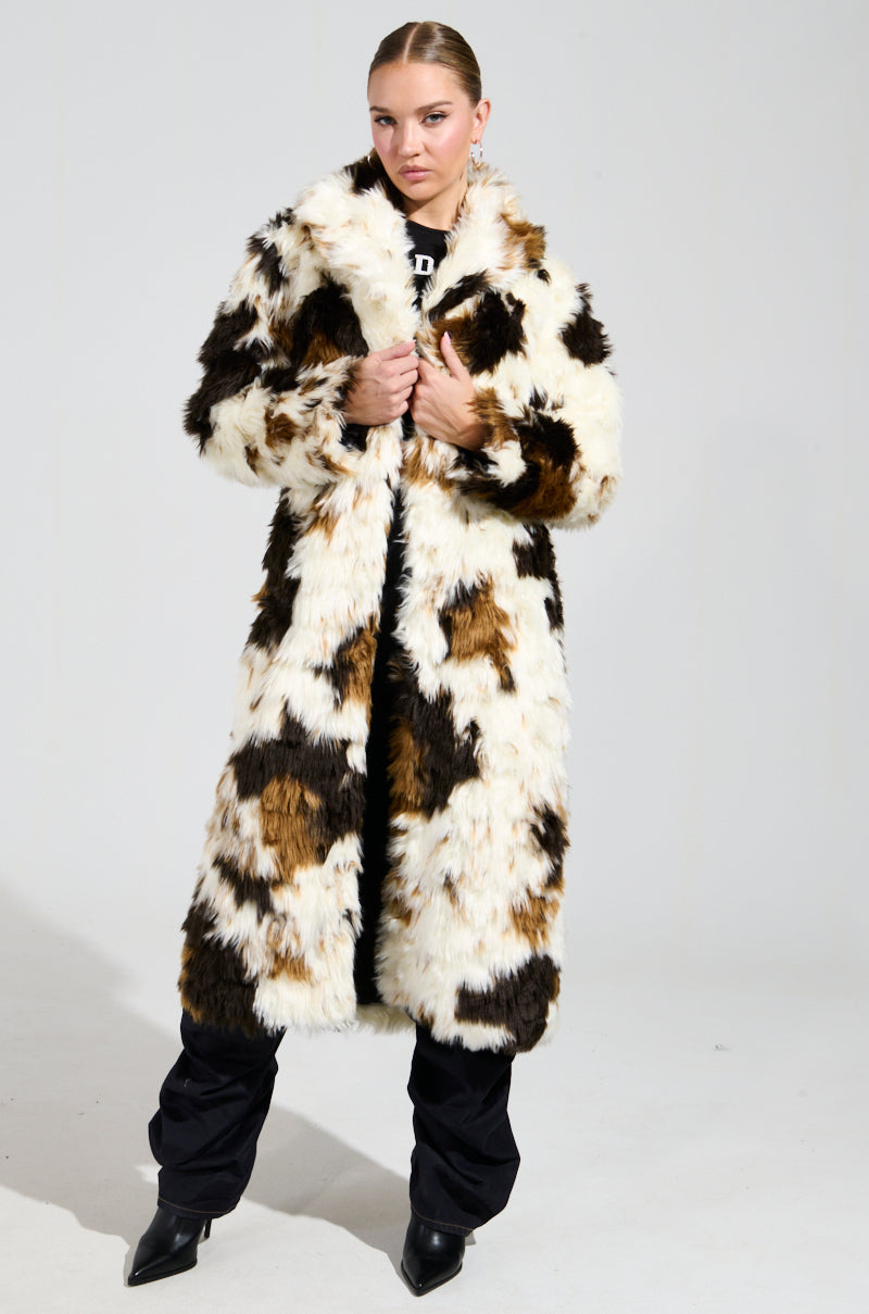 A person stands confidently, showcasing the stylish BRONX FAUX FUR COAT in a calico design of white, brown, and black patches. Their look is complemented by black pants and boots as they strike a pose against a simple backdrop.