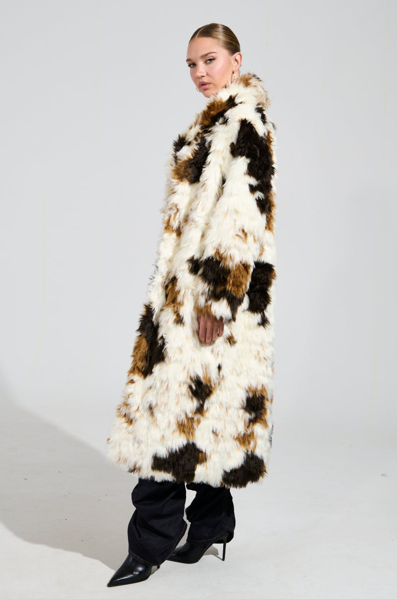 Wearing the BRONX FAUX FUR COAT in a calico colorway, a person stands sideways with slicked-back hair, paired with black pants and pointed shoes, posing against a plain, light background.