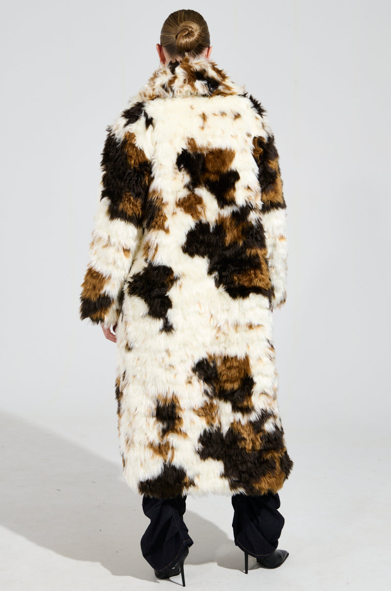 Wearing the BRONX FAUX FUR COAT, which showcases a shaggy-chic style in a calico colorway of brown, black, and white patchwork, a person appears with their hair neatly tied back, facing away from the camera against a plain, light-colored backdrop.