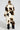 Wearing the BRONX FAUX FUR COAT, which showcases a shaggy-chic style in a calico colorway of brown, black, and white patchwork, a person appears with their hair neatly tied back, facing away from the camera against a plain, light-colored backdrop.