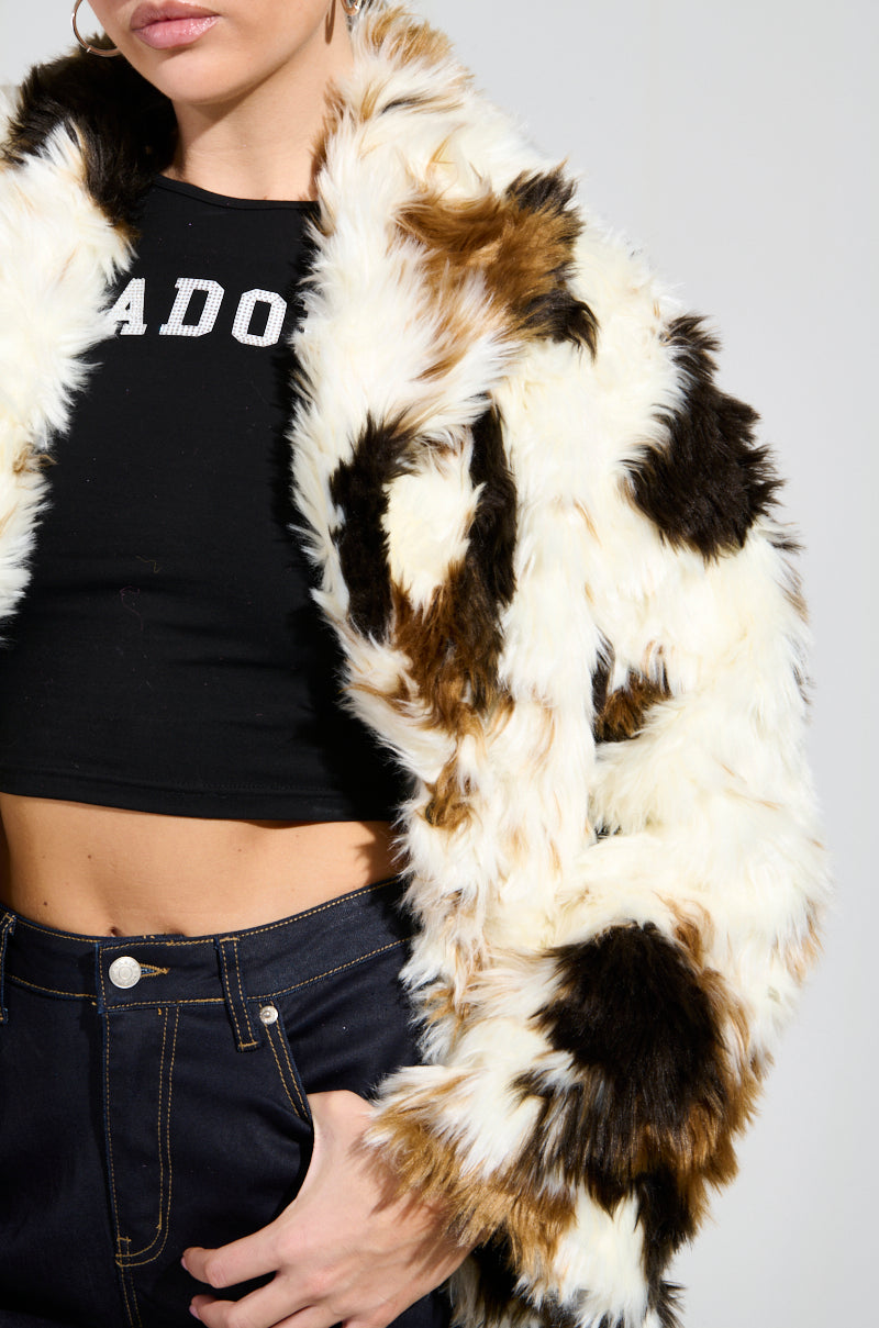 A person stuns in the BRONX FAUX FUR COAT, featuring a shaggy-chic, multicolored design, paired with a black crop top adorned with white lettering. The ensemble is completed with high-waisted dark jeans, keeping the focus on this fashion-forward outfit against a plain background.