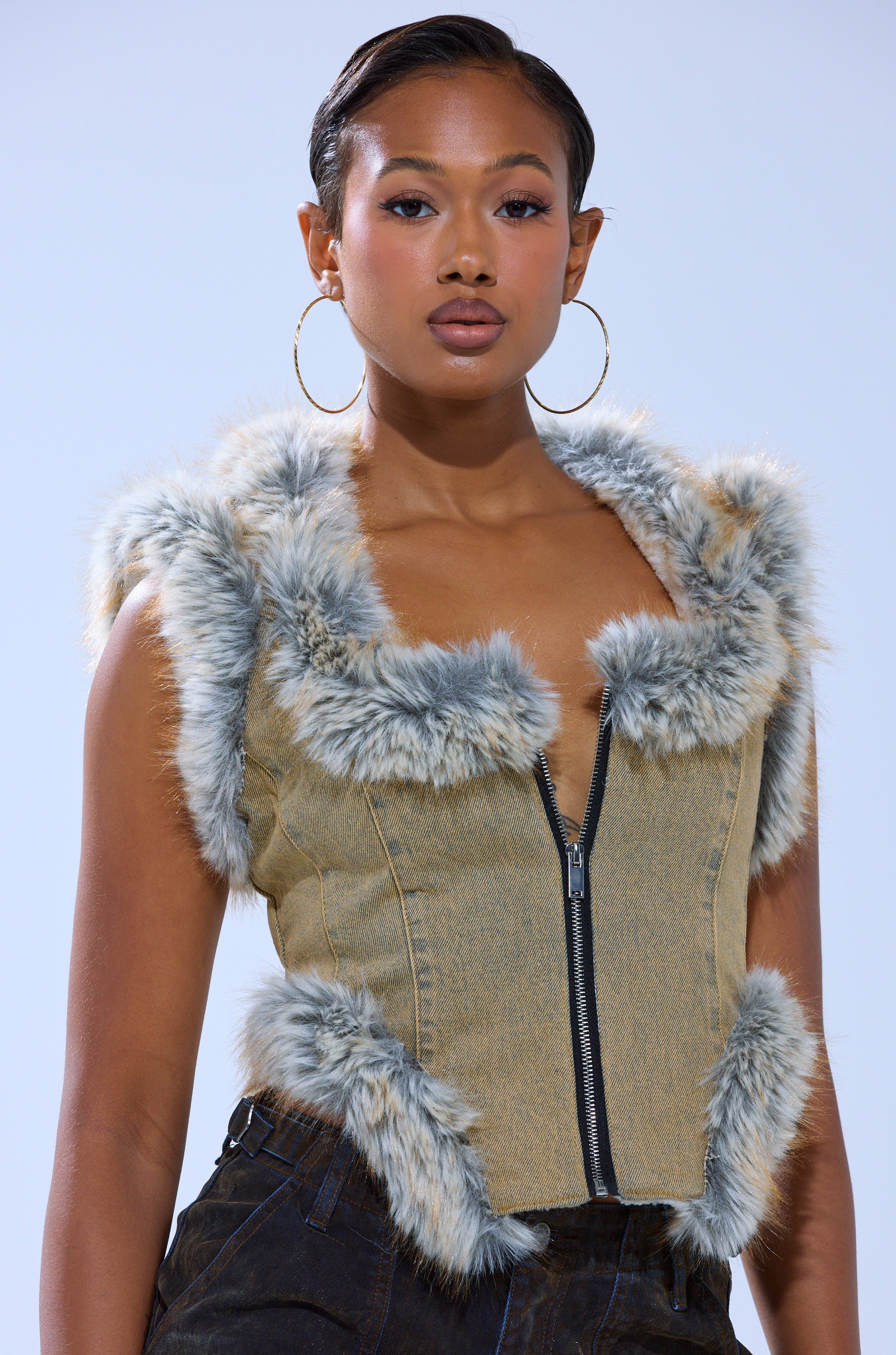 A woman poses confidently against a light blue background, wearing the ITTY BITTY FUR TRIM VEST—sleeveless with gray faux fur trim—and large hoop earrings.