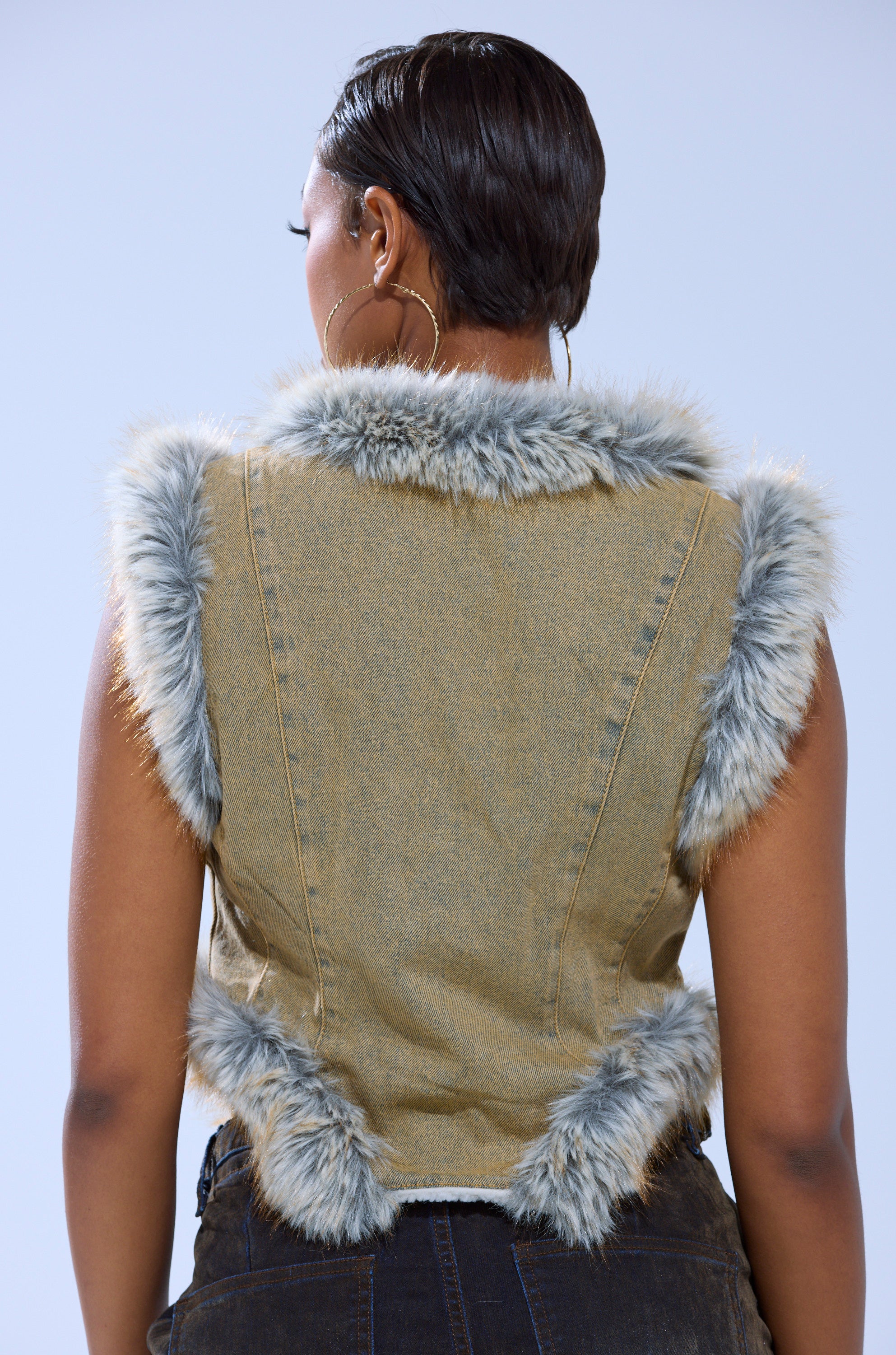 A person with short hair is seen from the back, wearing the ITTY BITTY FUR TRIM VEST—a sleeveless denim vest with light gray faux fur along the shoulders, armholes, and hem, featuring a corset-inspired cut and paired with dark pants.