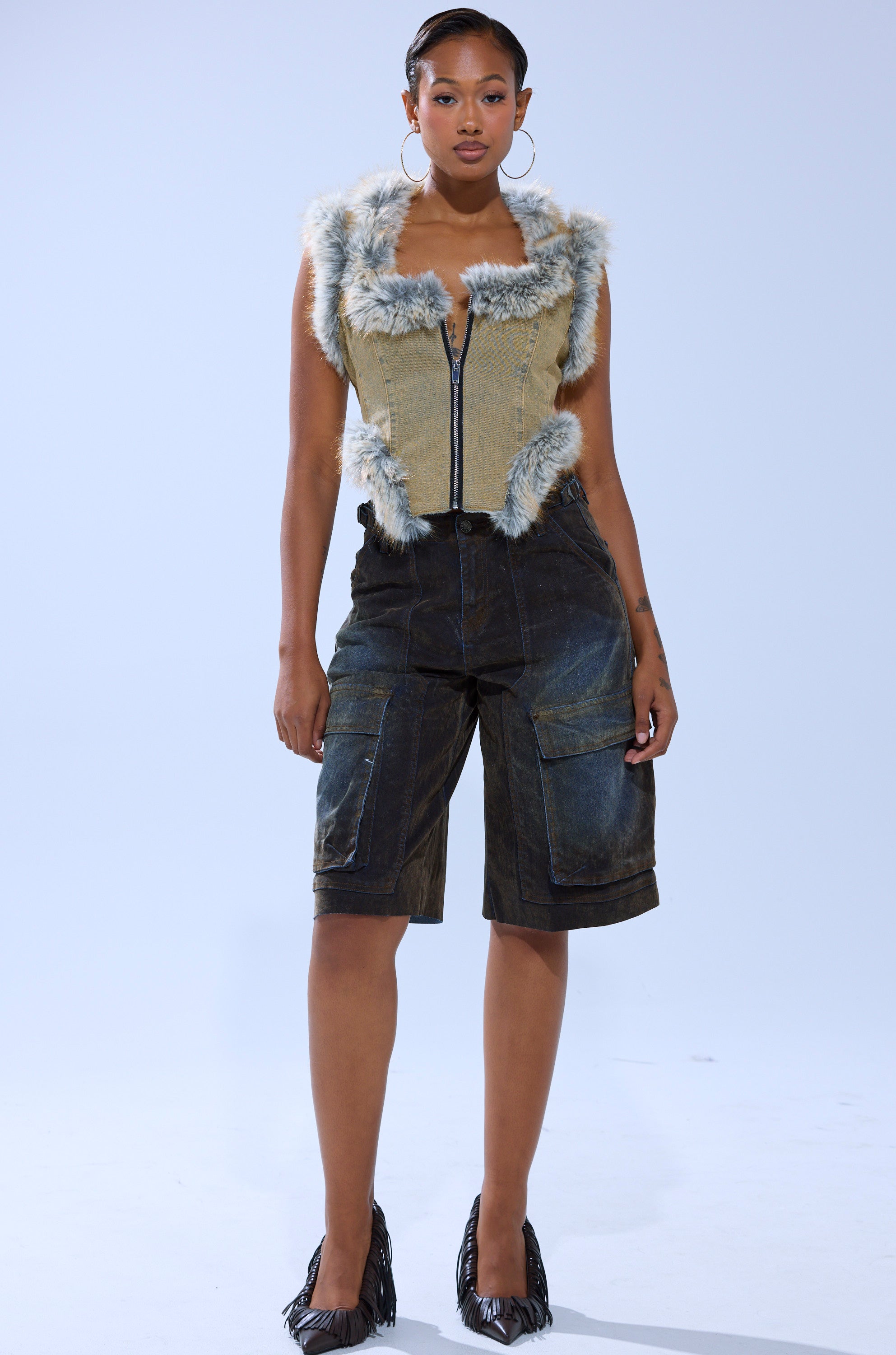 A woman exudes confidence in the ITTY BITTY FUR TRIM VEST with a corset-inspired hem, paired with knee-length dark denim shorts, hoop earrings, and black heels. Her hair is pulled back against a plain light gray background.
