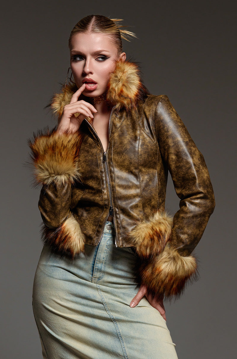The individual poses confidently, highlighting the DOWN BAD FUR TRIM MOTO—a vintage-style cropped faux leather jacket accented with fluffy fur trim. They have one hand on their chin and are paired with a long denim skirt, set against a neutral background.