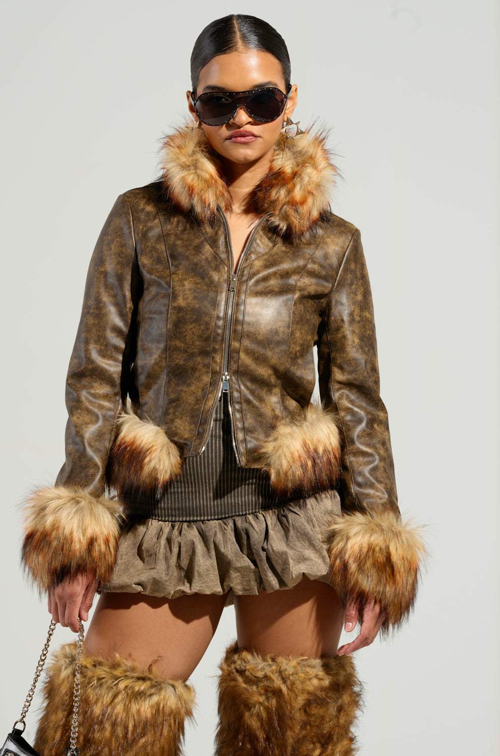 A person dressed in the DOWN BAD FUR TRIM MOTO, featuring a brown faux leather jacket with fur trim, paired with a matching cropped silhouette skirt and fur boots. They complete the ensemble with sunglasses and a purse with a chain strap, presenting an elegant blend of textures in earthy tones.