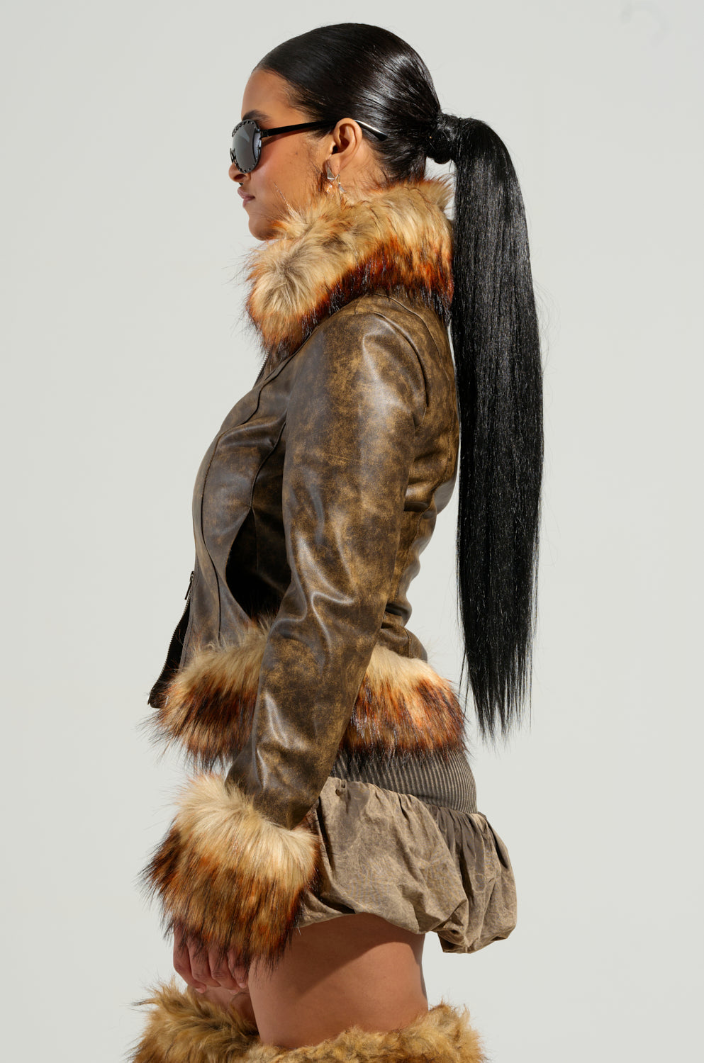 A woman showcases the DOWN BAD FUR TRIM MOTO, a distressed faux leather jacket boasting a cropped silhouette with luxurious fur trim on the collar and cuffs. It pairs perfectly with a matching brown skirt and fur boots, while her sleek ponytail and dark sunglasses add the finishing touches to this chic ensemble.