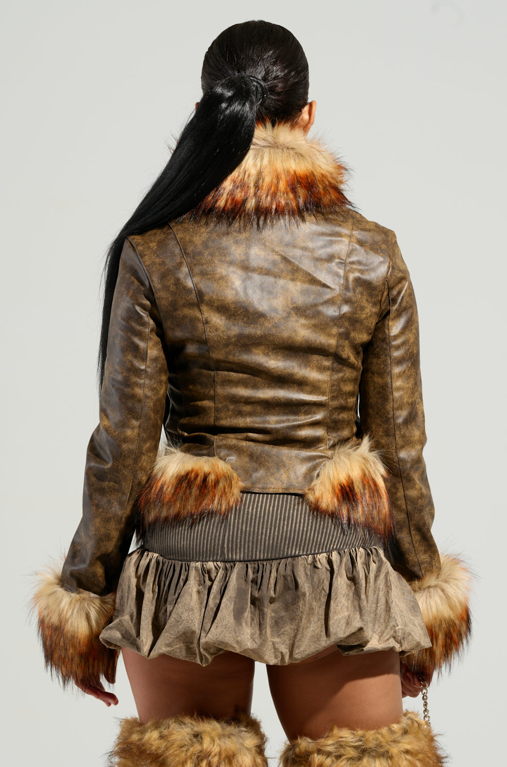 A person with long dark hair is wearing the DOWN BAD FUR TRIM MOTO, a brown faux leather jacket with fur trim. They are standing with their back to the camera, highlighting a chic cropped silhouette paired with a matching skirt and fur-trimmed boots.