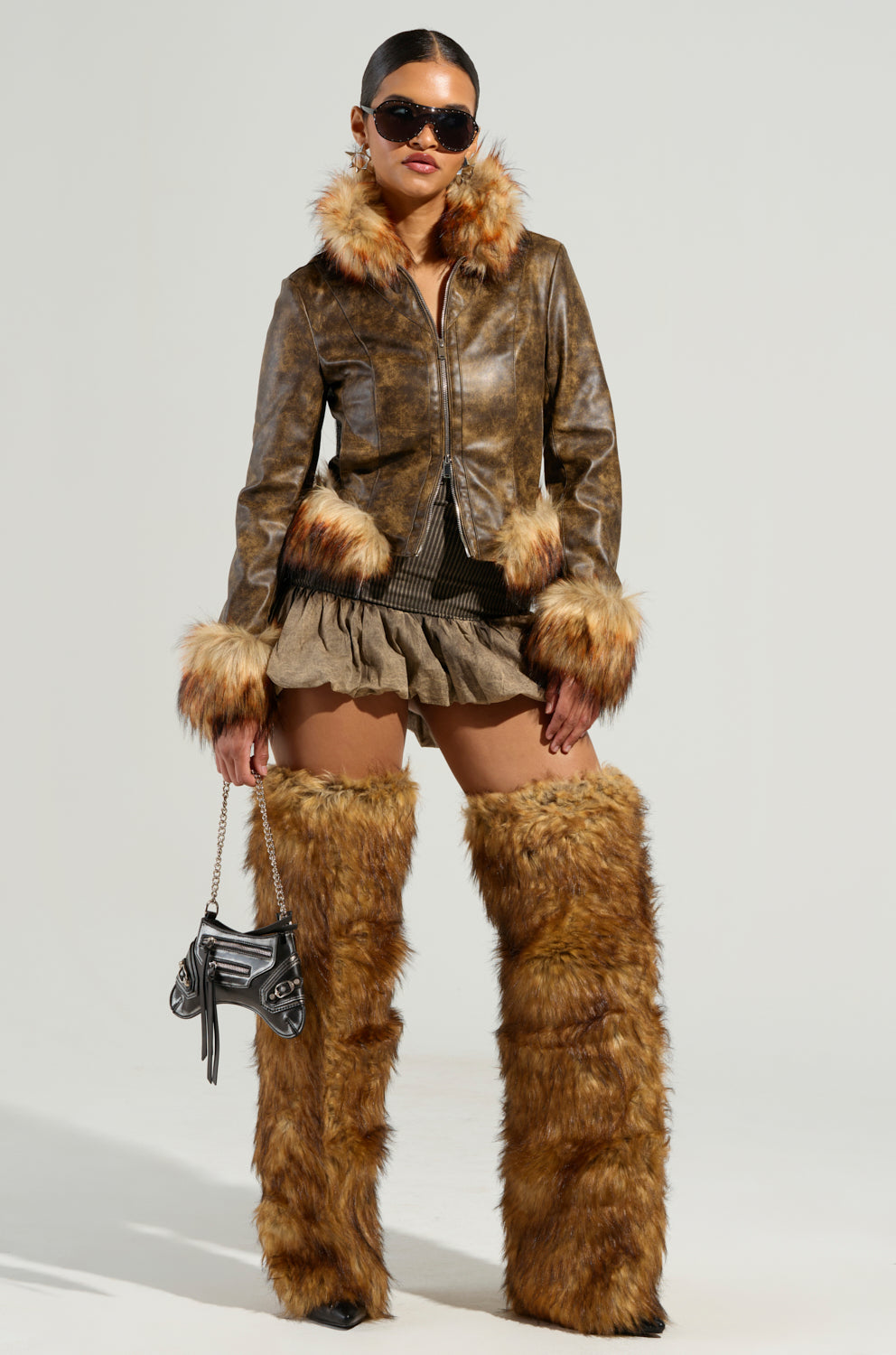 A fashionable person exudes confidence in sunglasses and the DOWN BAD FUR TRIM MOTO faux leather cropped silhouette jacket. Complemented by fur shorts, tall fur boots, and a small handbag, they make a striking impression against a neutral background.