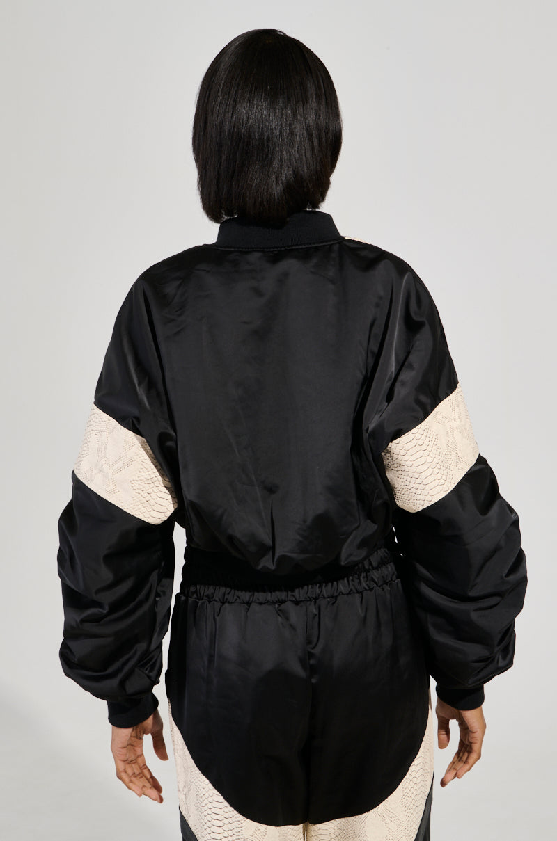 A person with shoulder-length black hair is shown from behind, wearing the LAINA LIGHTWEIGHT BOMBER, which features bold snakeskin-print stripes on the sleeves. The jacket has a loose fit, set against a plain gray background.
