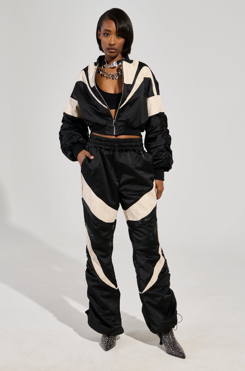 A person confidently stands with hands in pockets, wearing the LAINA LIGHTWEIGHT BOMBER, a stylish black and white tracksuit featuring geometric patterns and color block panels. They sport short hair, a layered necklace, and patterned shoes against a plain background.