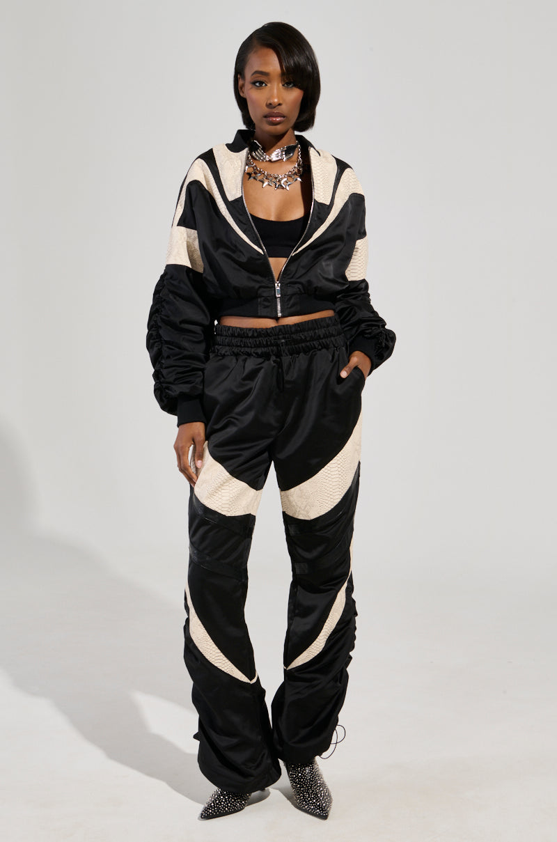 Wearing the LAINA LIGHTWEIGHT BOMBER, a person exudes confidence in a chic black and beige tracksuit with a cropped jacket and high-waisted pants accented by subtle snakeskin print panels. They complete the look with a silver choker, casually placing one hand in their pocket against a plain gray backdrop.