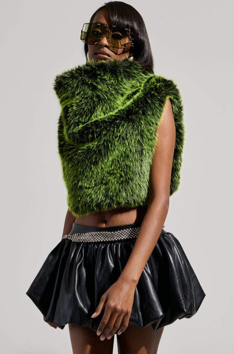 Standing against a neutral background, a person exudes confidence in an eye-catching ensemble featuring the "HEADED TO ASPEN SLEEVELESS FUR TOP IN GREEN" paired with a black leather skirt in a cropped silhouette. Complemented by oversized sunglasses and a chain belt, the look is undeniably bold and fashionable.