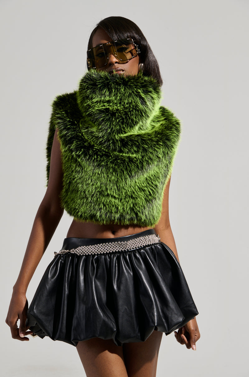 Wearing the "Headed to Aspen Sleeveless Fur Top in Green," this person with long hair complements their oversized sunglasses and vibrant top with a short black leather skirt and a silver chain belt. The cropped silhouette stands out against the plain background, highlighting the stylish outfit.