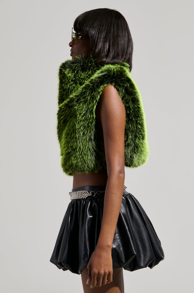 A person in profile wears sunglasses, the HEADED TO ASPEN SLEEVELESS FUR TOP IN GREEN, and a short black leather skirt with a chain belt. The cropped silhouette stands out against the plain gray background, emphasizing the striking ensemble.