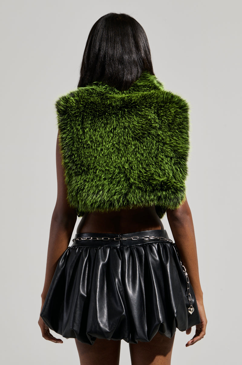 A person with long black hair stands facing away, wearing the HEADED TO ASPEN SLEEVELESS FUR TOP IN GREEN and a short black leather skirt with a chain belt. The plain gray backdrop accentuates the striking silhouette.