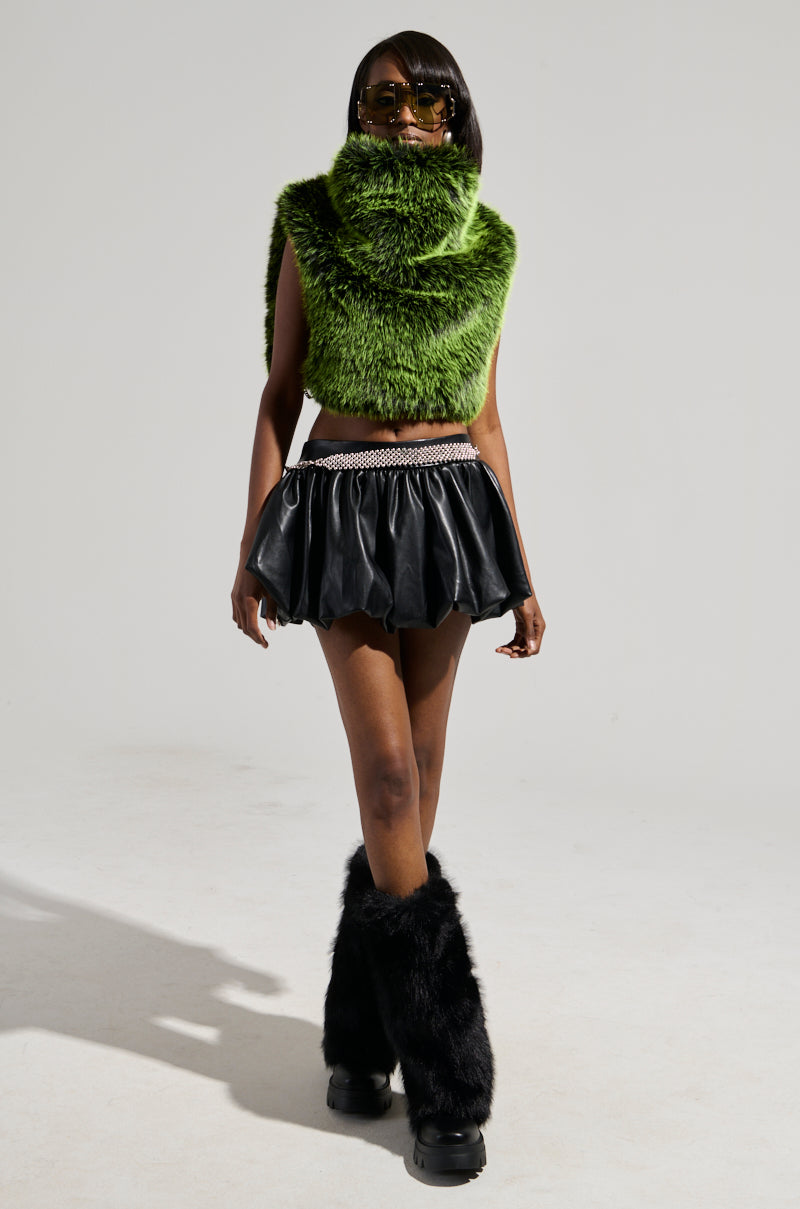 A person stands confidently, wearing the HEADED TO ASPEN SLEEVELESS FUR TOP IN GREEN that showcases a chic, cropped silhouette. Paired with sunglasses, a black mini skirt with a studded belt, and furry boots, the vibrant ensemble pops against the plain background.