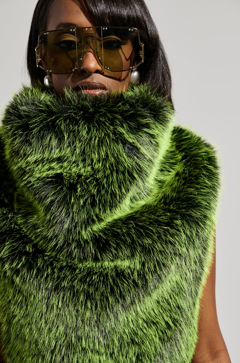 A person wearing large sunglasses and pearl earrings is elegantly draped in the luxurious HEADED TO ASPEN SLEEVELESS FUR TOP IN GREEN, boasting a vibrant color and cropped silhouette, set against a plain background.
