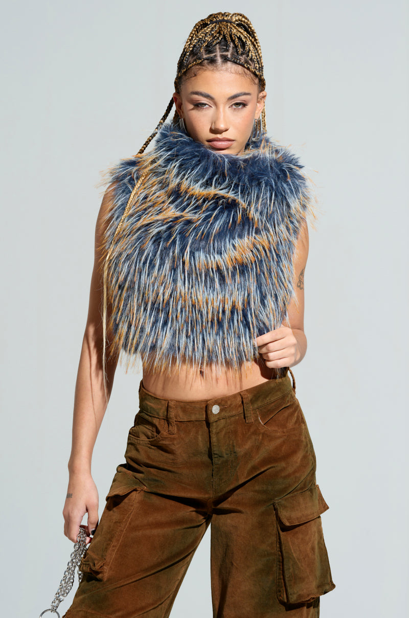 A person with braided hair poses confidently in the HEADED TO ASPEN SLEEVELESS FUR TOP IN BLUE, paired with brown cargo pants. They hold a chain accessory in one hand, standing against a plain light gray background.