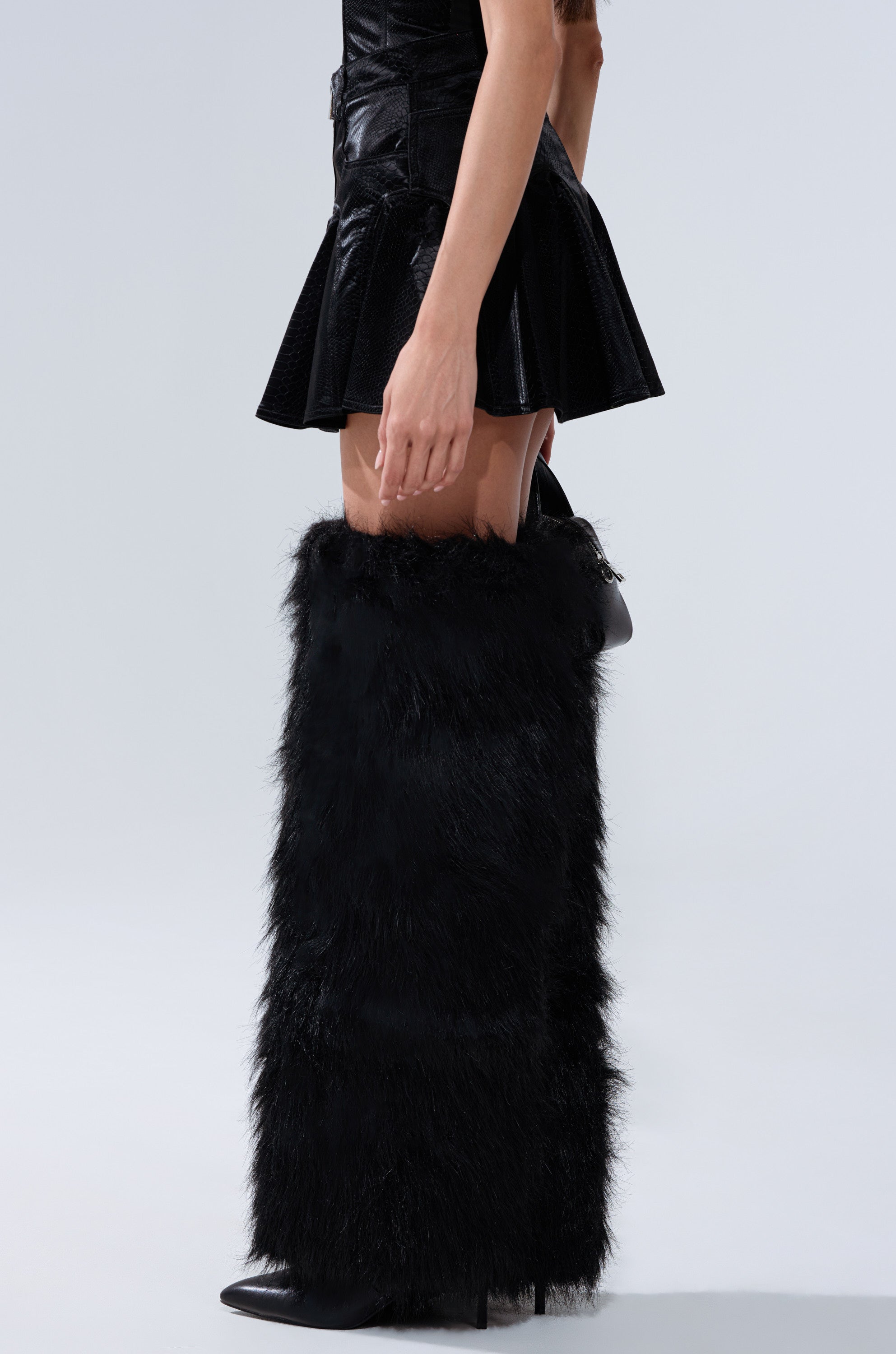Wearing the SWEETIE PIE MINI SKIRT in black faux leather with tall, fluffy black faux fur boots over the knees, a person stands against a plain light gray background.
