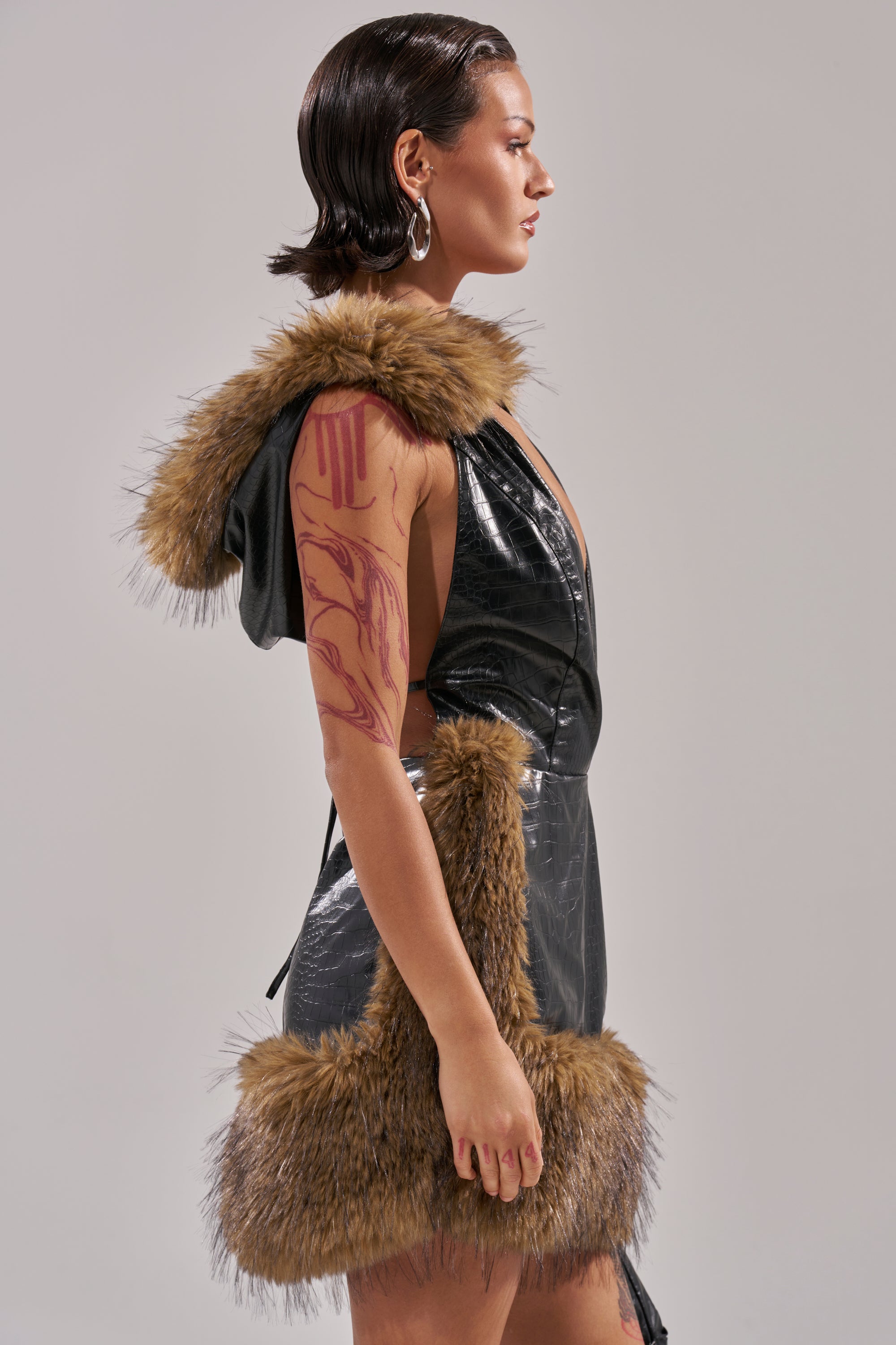 A model in profile wears the ATE DOWN PU MINI DRESS—a shiny black sleeveless hooded mini dress with camel faux fur trim—showing a large red abstract arm tattoo and silver hoop earrings, set against a plain gray backdrop.