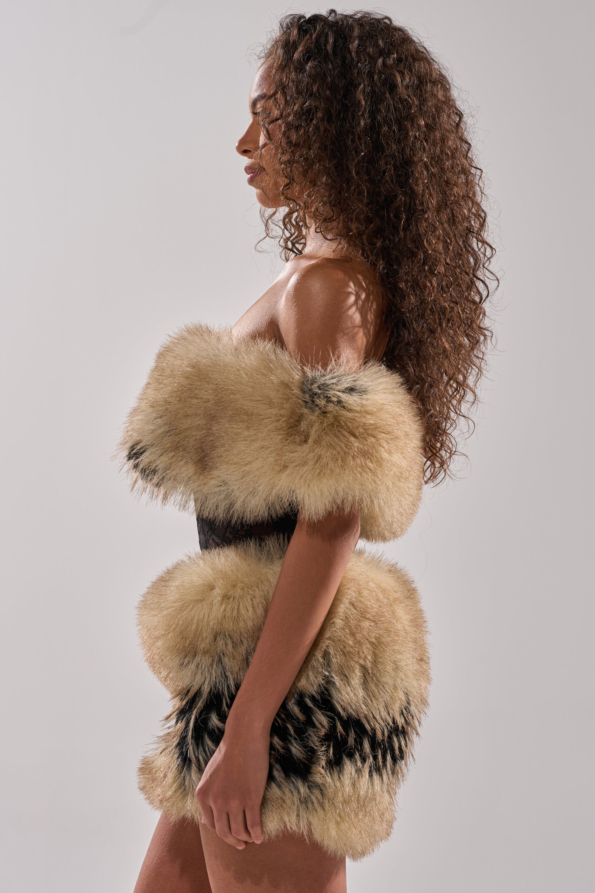 A woman with long curly hair stands in profile, wearing the COOL GIRLS ONLY LACE FUR MINI DRESS in tan and black. The simple, light background highlights the unique textures of her strapless mini dress.