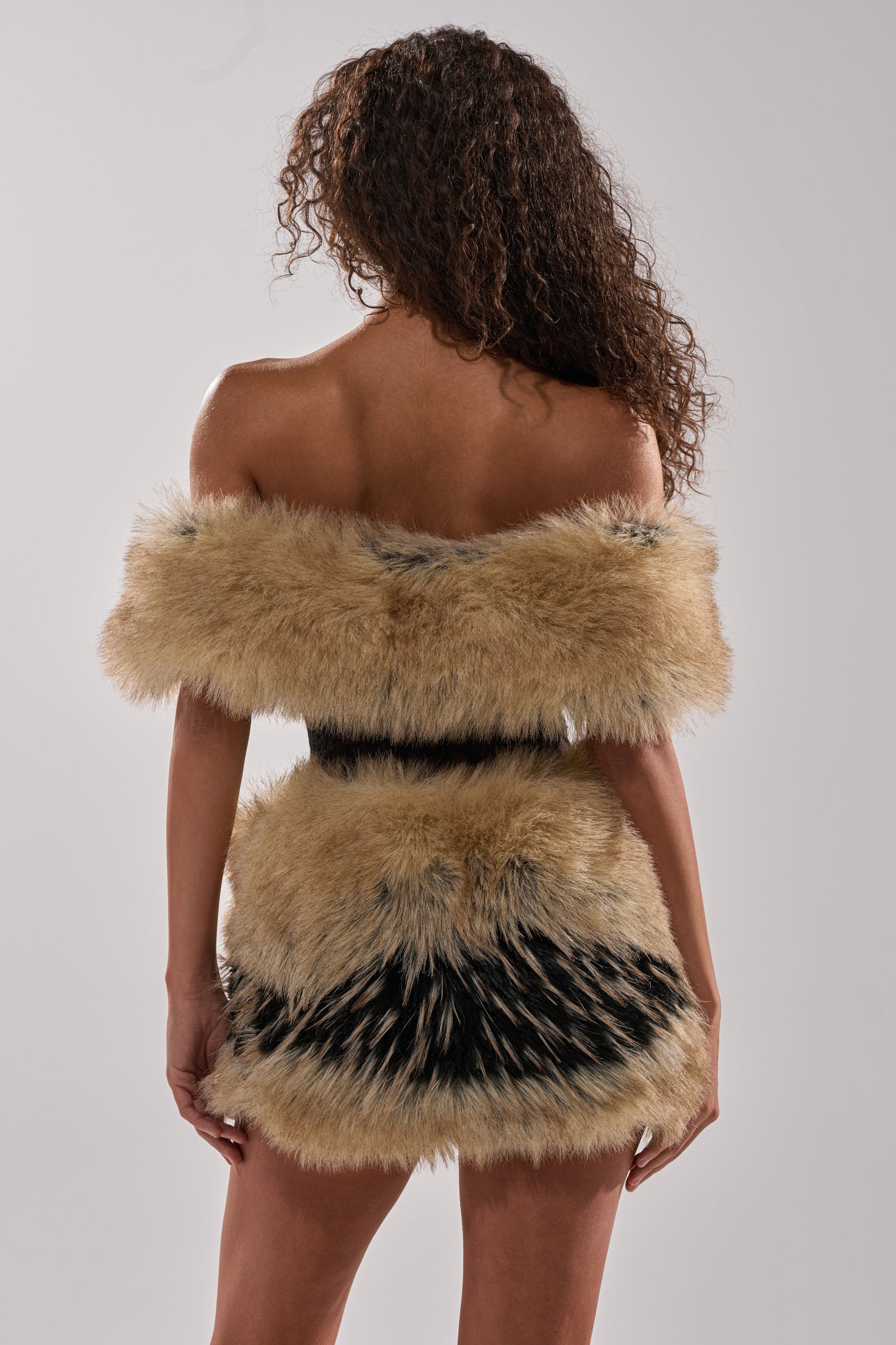 A woman with curly hair, seen from the back, wears the COOL GIRLS ONLY LACE FUR MINI DRESS in tan and black off-the-shoulder style, standing against a plain light gray background.