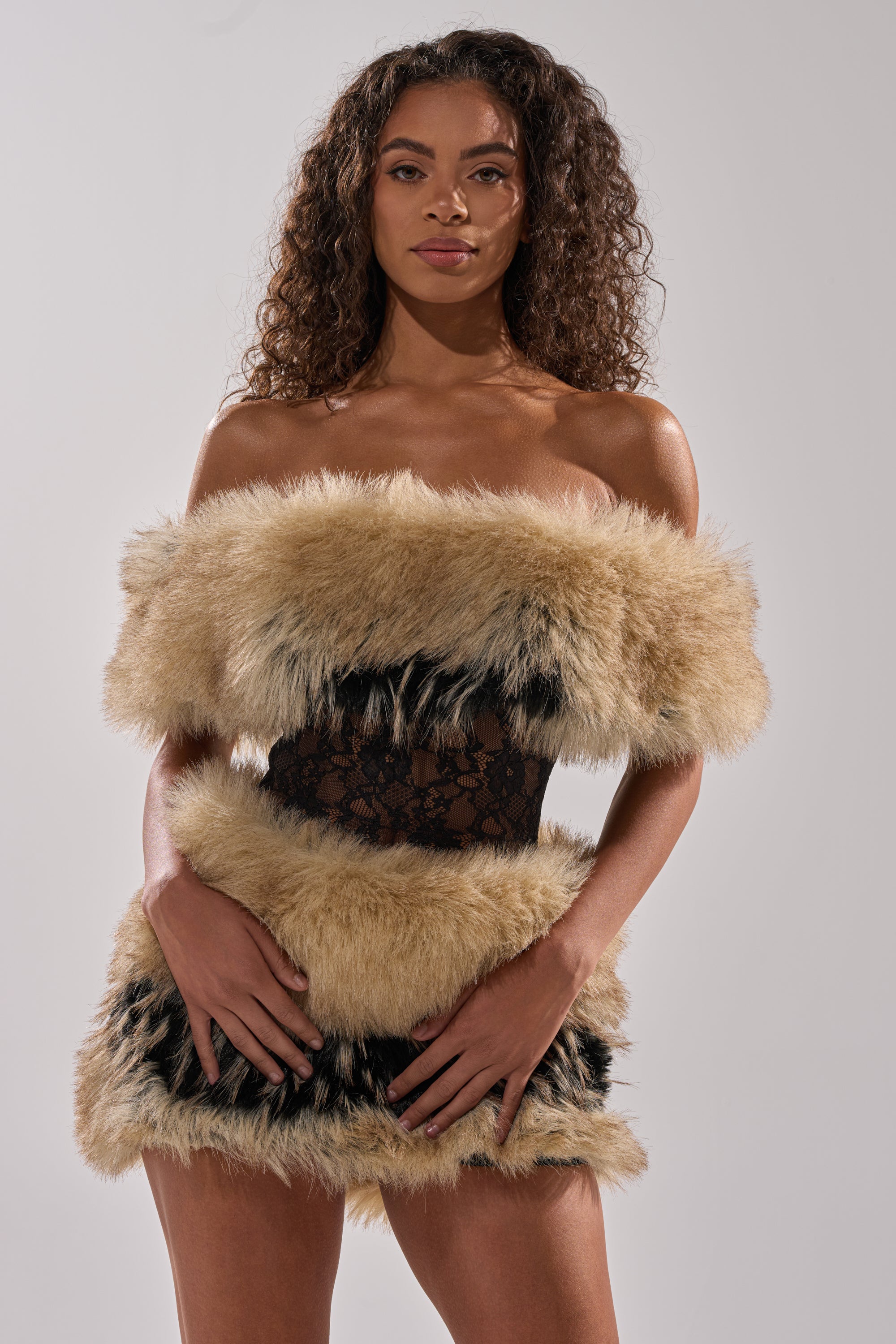 A woman with curly hair poses confidently in the COOL GIRLS ONLY LACE FUR MINI DRESS, showcasing black lace and light brown faux fur panels, set against a plain light gray background.