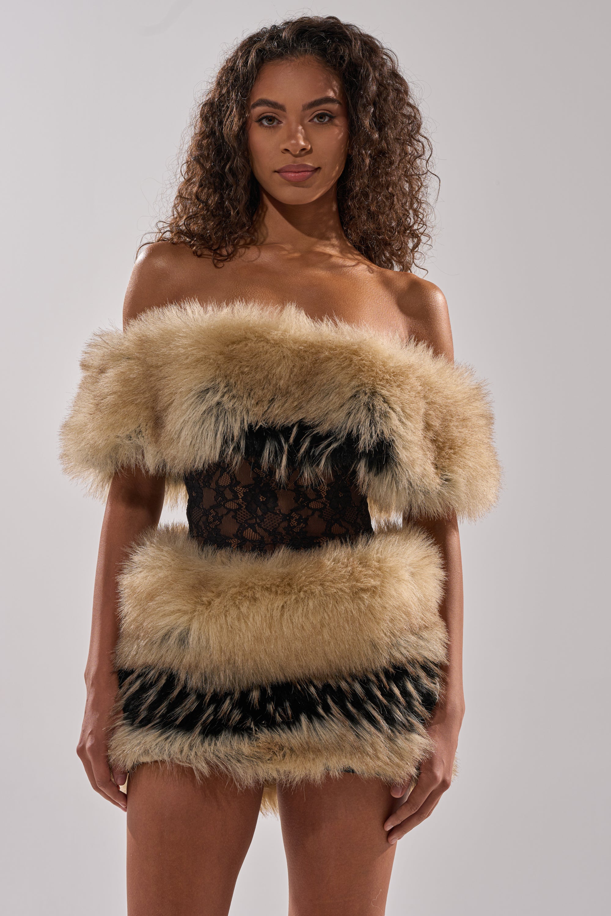 A woman with curly hair models the COOL GIRLS ONLY LACE FUR MINI DRESS, an off-shoulder beige and black faux fur mini dress with a central black lace panel, set against a plain light background.