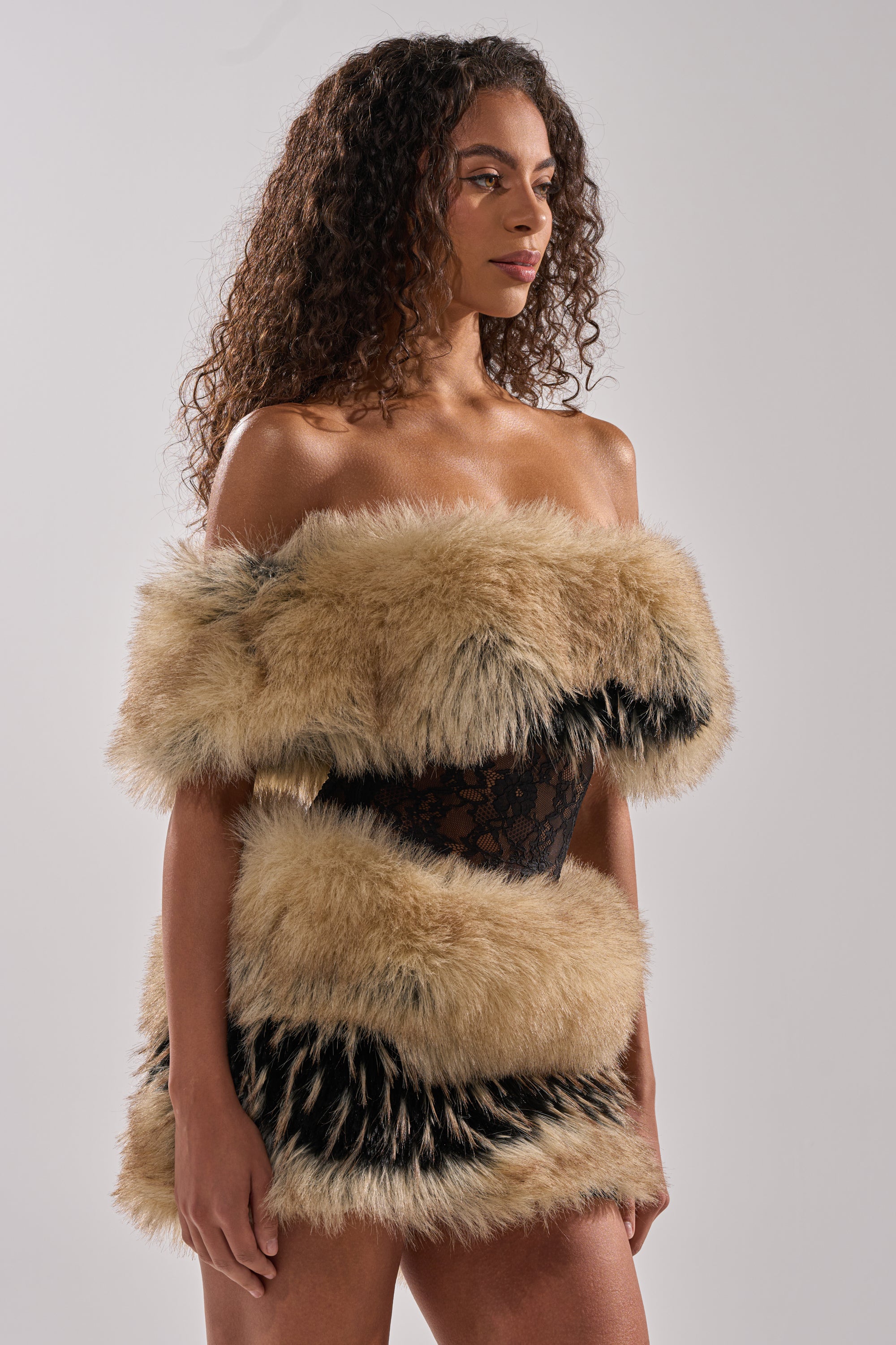A woman with curly hair wears the COOL GIRLS ONLY LACE FUR MINI DRESS, a sleeveless mini with faux fur trim and bold tan and black bands, standing against a plain light gray background.