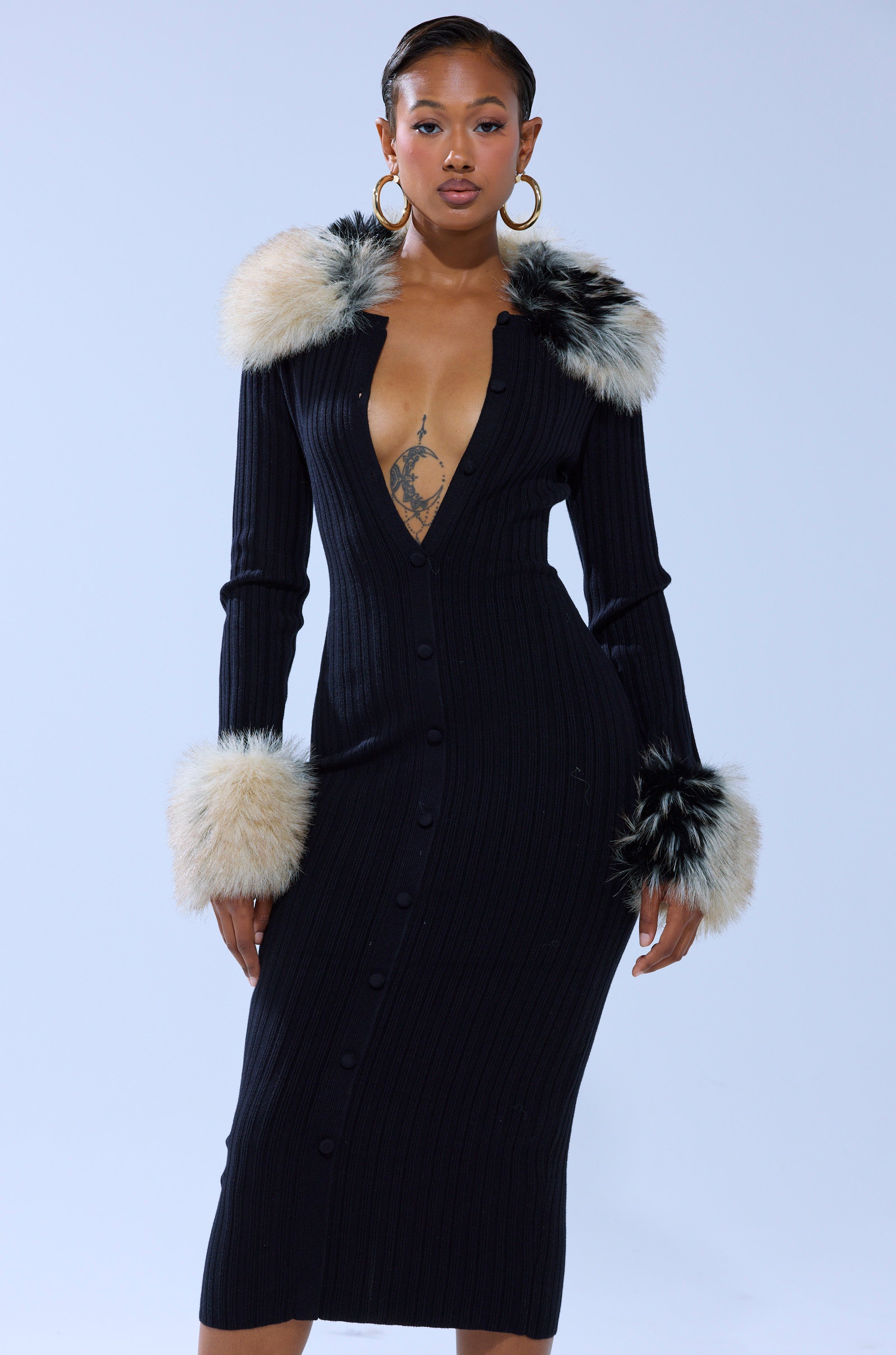 A woman confidently poses in the MICHELLE KNIT MIDI DRESS WITH FUR ACCENTS, featuring a ribbed knit, deep V-neckline, faux fur collar and cuffs, paired with large hoop earrings against a light background.