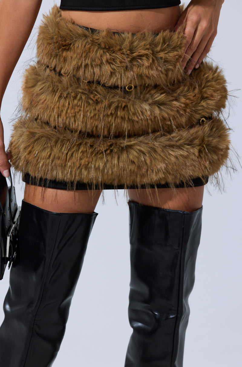 A person models the WILD AND FREE FUR MINI SKIRT—a golden-brown faux fur mini skirt with layered tiers—paired with thigh-high black leather boots. Only the lower torso and legs are visible against a plain background. 1