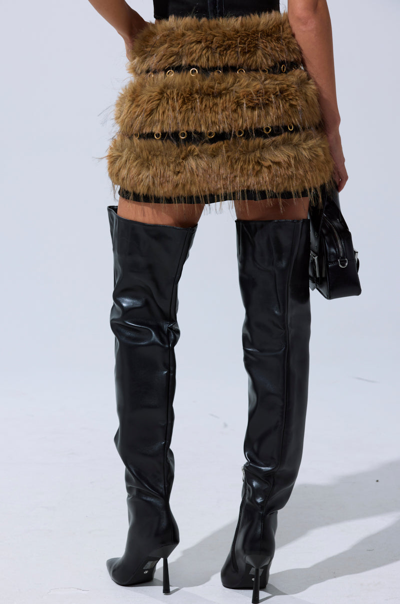 Wearing the WILD AND FREE FUR MINI SKIRT in golden-brown with black grommet details and thigh-high black leather stiletto boots, a person stands against a plain light background. 5
