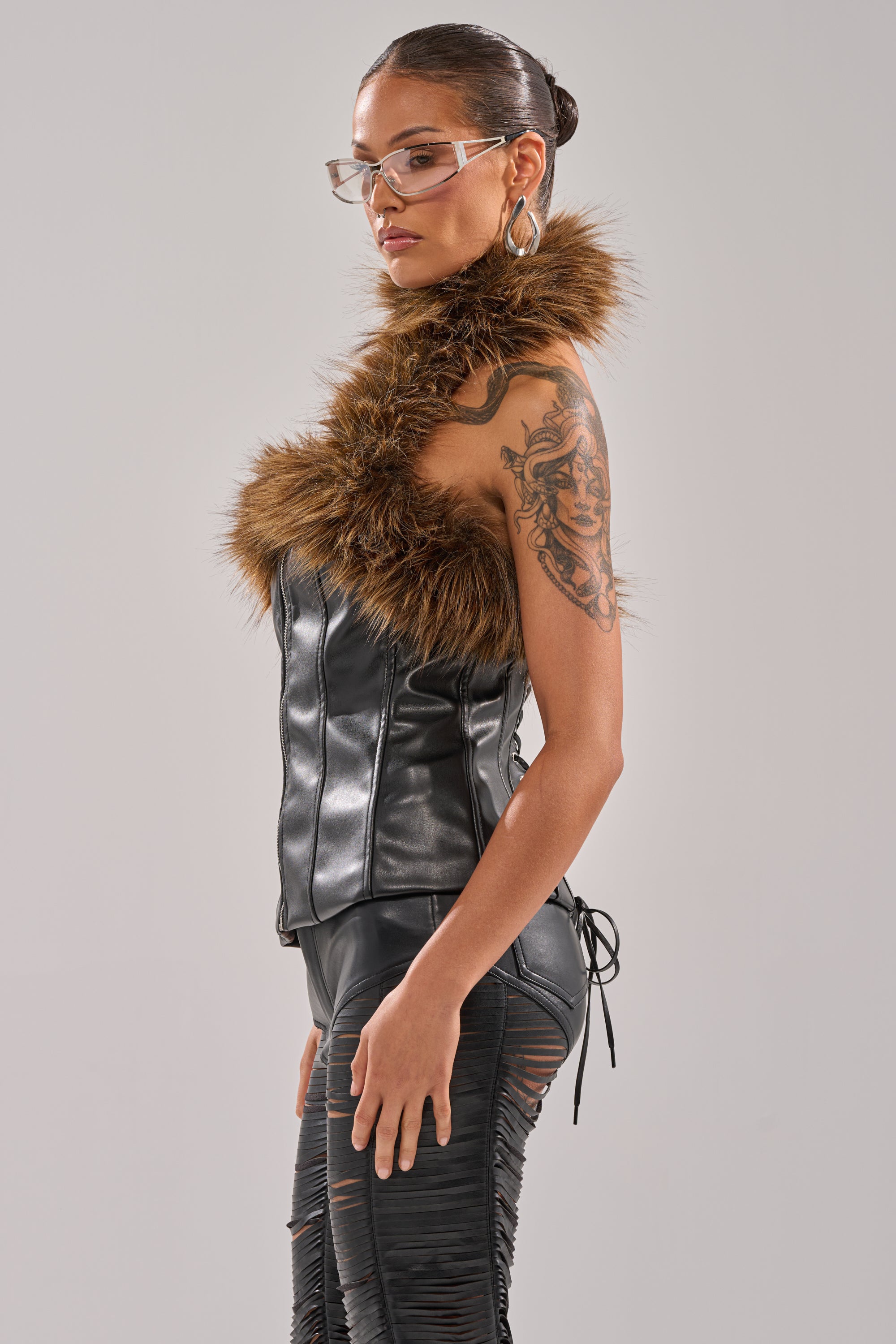 A woman in clear glasses stands sideways, her sleek bun highlighting an upper arm tattoo. She wears the bold ARE YOU FOR REAL FUR TRIM CORSET paired with black ripped pants, adding edgy flair to her modern look. 4