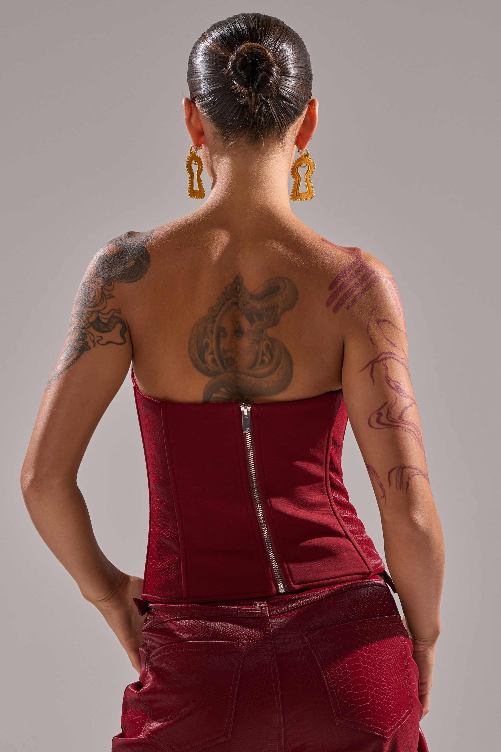 A person with back and arm tattoos faces away, hair in a neat bun, wearing the TOO HOT FOR YOU FAUX LEATHER CORSET in maroon with a zipper, maroon pants, and large statement earrings.