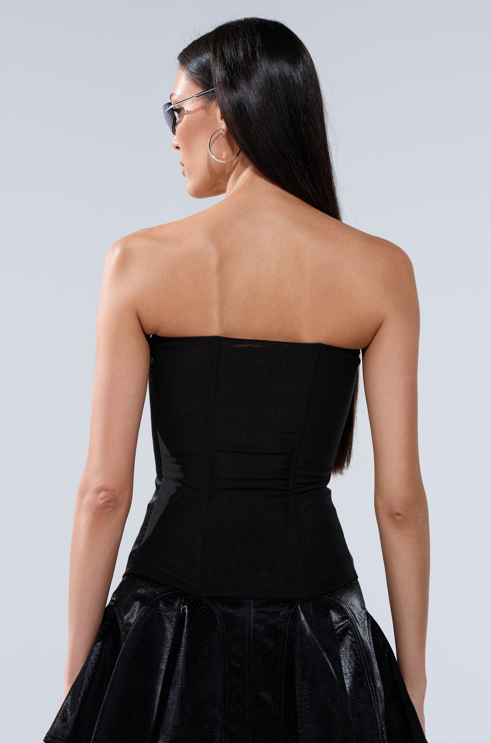 A woman with long, straight dark hair is seen from behind wearing the SWEETIE PIE CORSET and a shiny black textured skirt, paired with hoop earrings and sunglasses. The background is plain and light-colored. 7