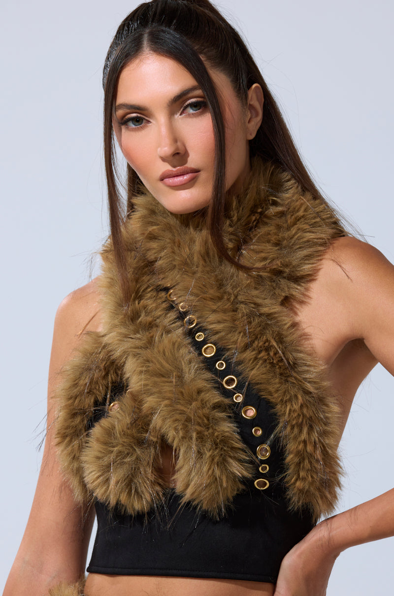 A woman with long, straight brown hair wears the WILD AND FREE FUR BLOUSE—a statement piece featuring fluffy brown fur and gold-tone grommet details—over a black crop top, posing against a plain light background. 1