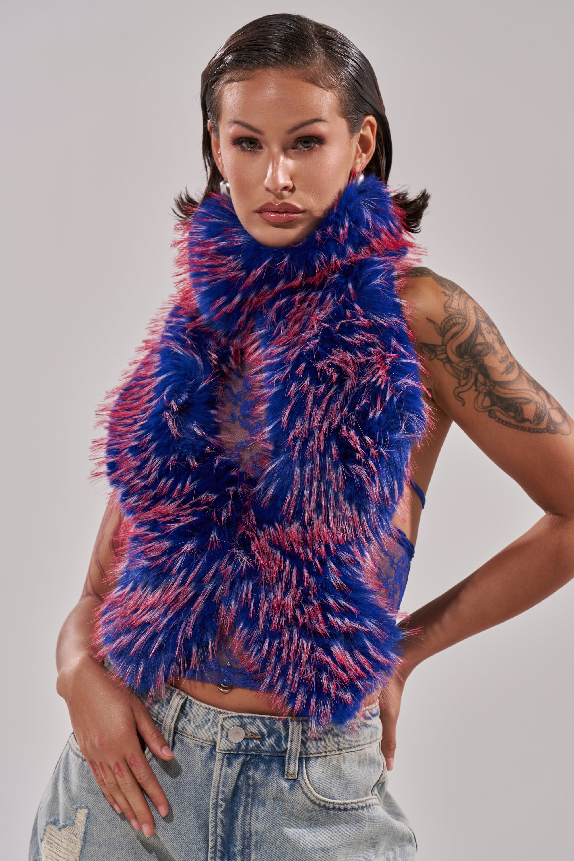 A woman with slicked-back hair models the FEEL ME BABY FUR BLOUSE IN BLUE, a fluffy blue and pink top. She shows her tattooed arm and poses confidently in distressed light blue jeans against a plain gray background. 2