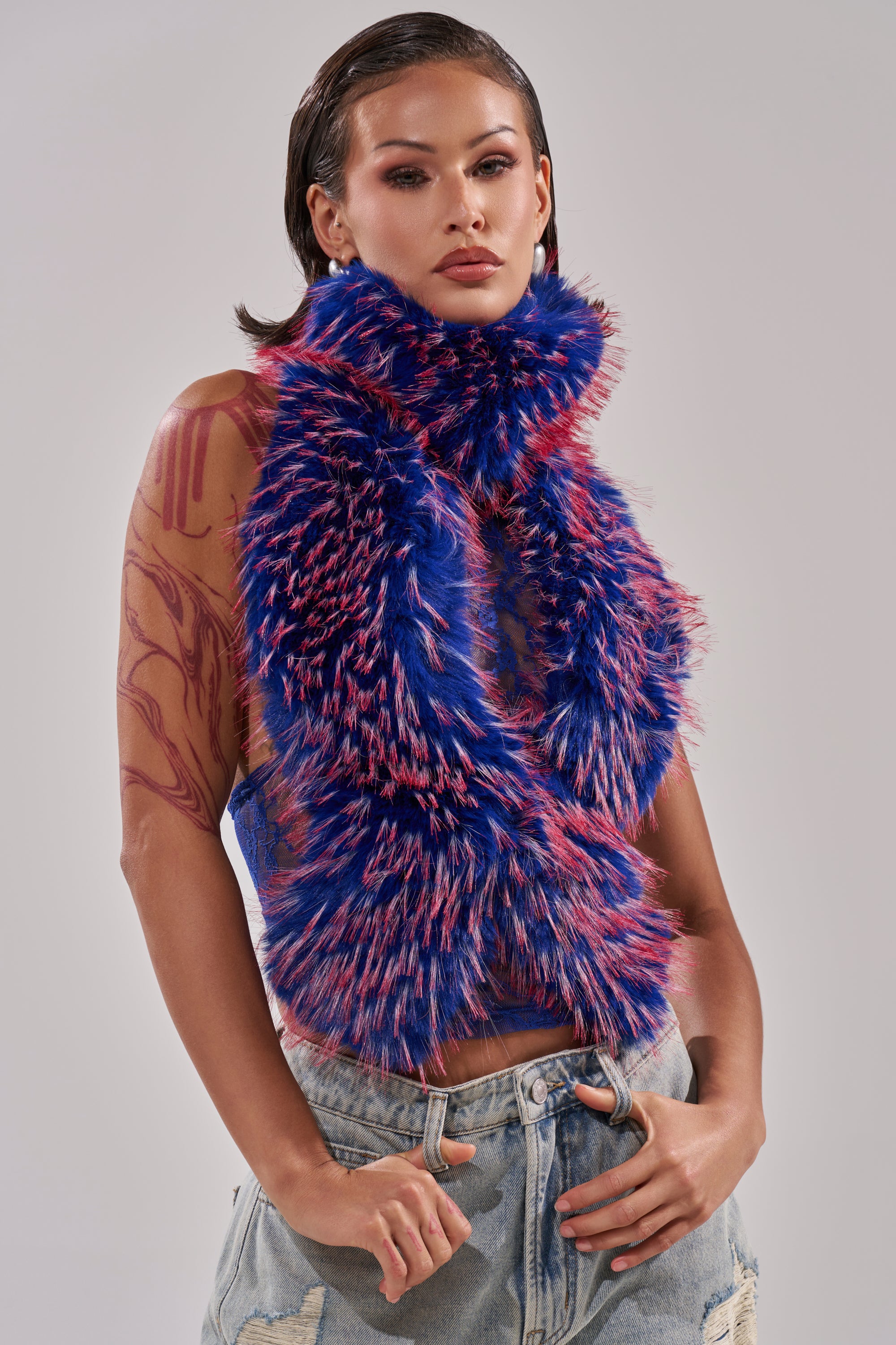 A woman with wet-look hair wears the FEEL ME BABY FUR BLOUSE IN BLUE over a sheer top with red abstract designs and distressed denim jeans, standing against a light gray background. 3