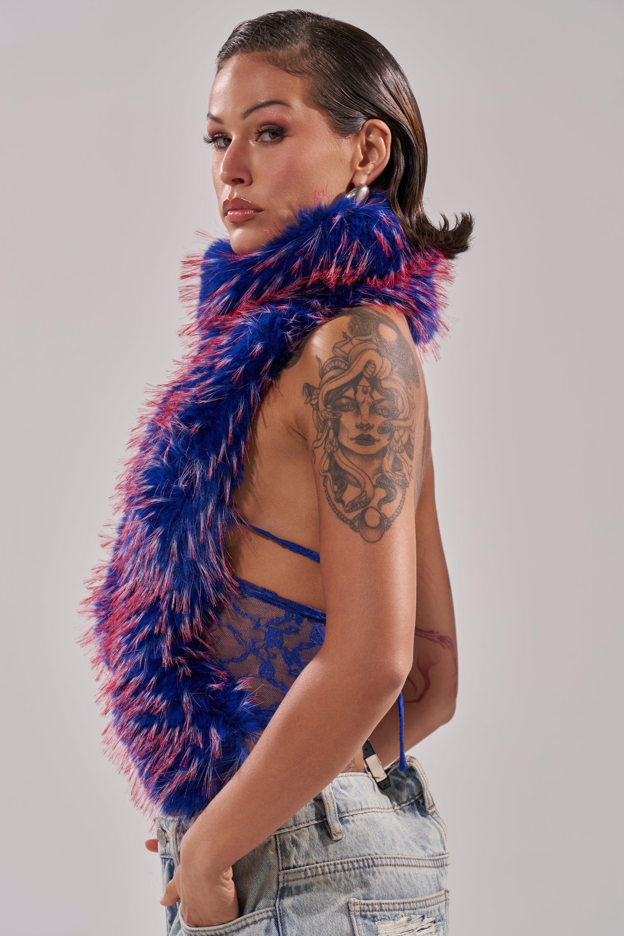 A person with slicked-back hair wears a blue FEEL ME BABY FUR BLOUSE, a blue lace top, and light denim jeans. Their upper arm features a tattoo of a woman's face and headdress. The background is plain light gray. 4