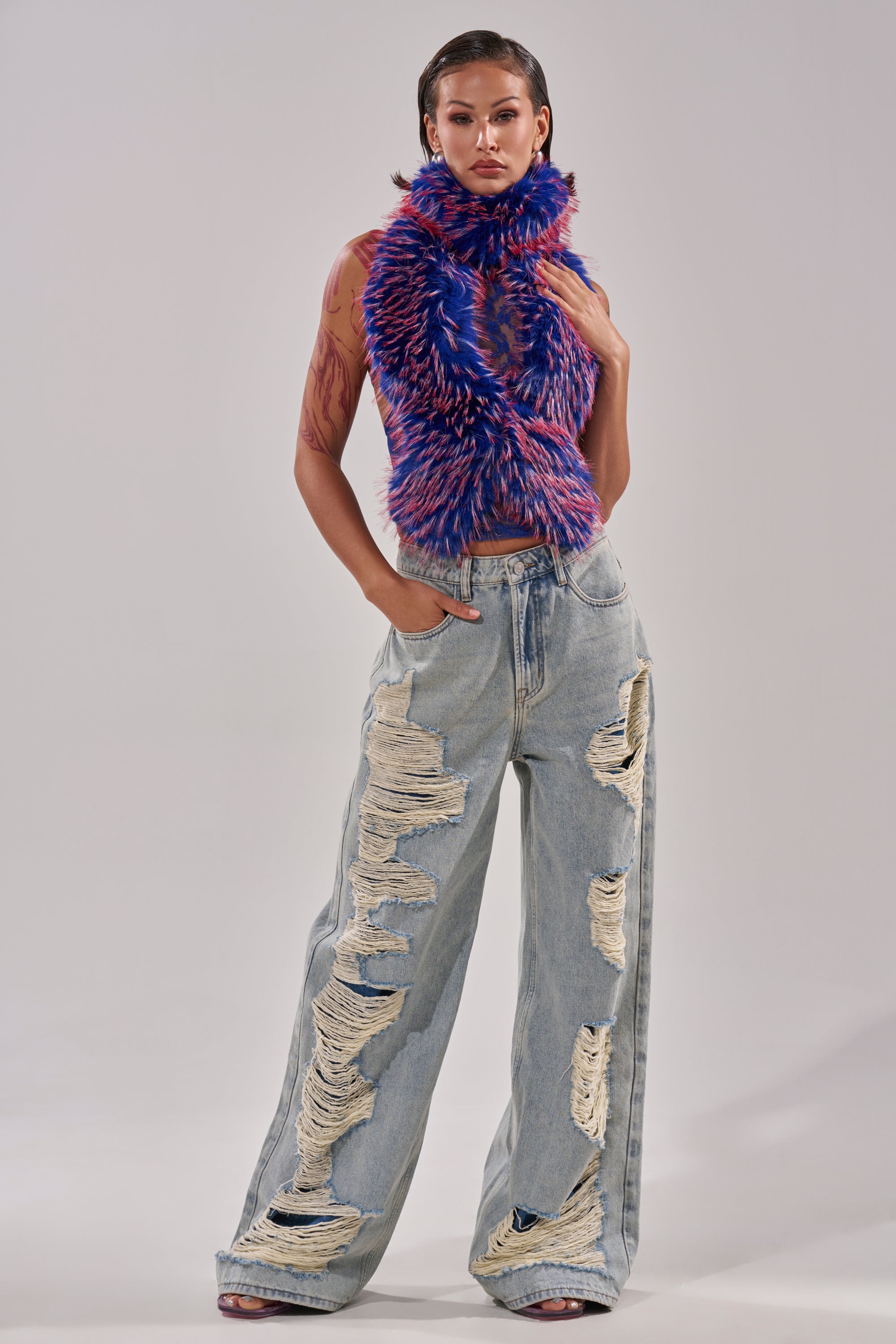 A woman stands confidently in the FEEL ME BABY FUR BLOUSE IN BLUE—a sleeveless, textured faux fur crop top—paired with light blue, wide-leg ripped jeans, posing with one hand in her pocket against a plain background. 6