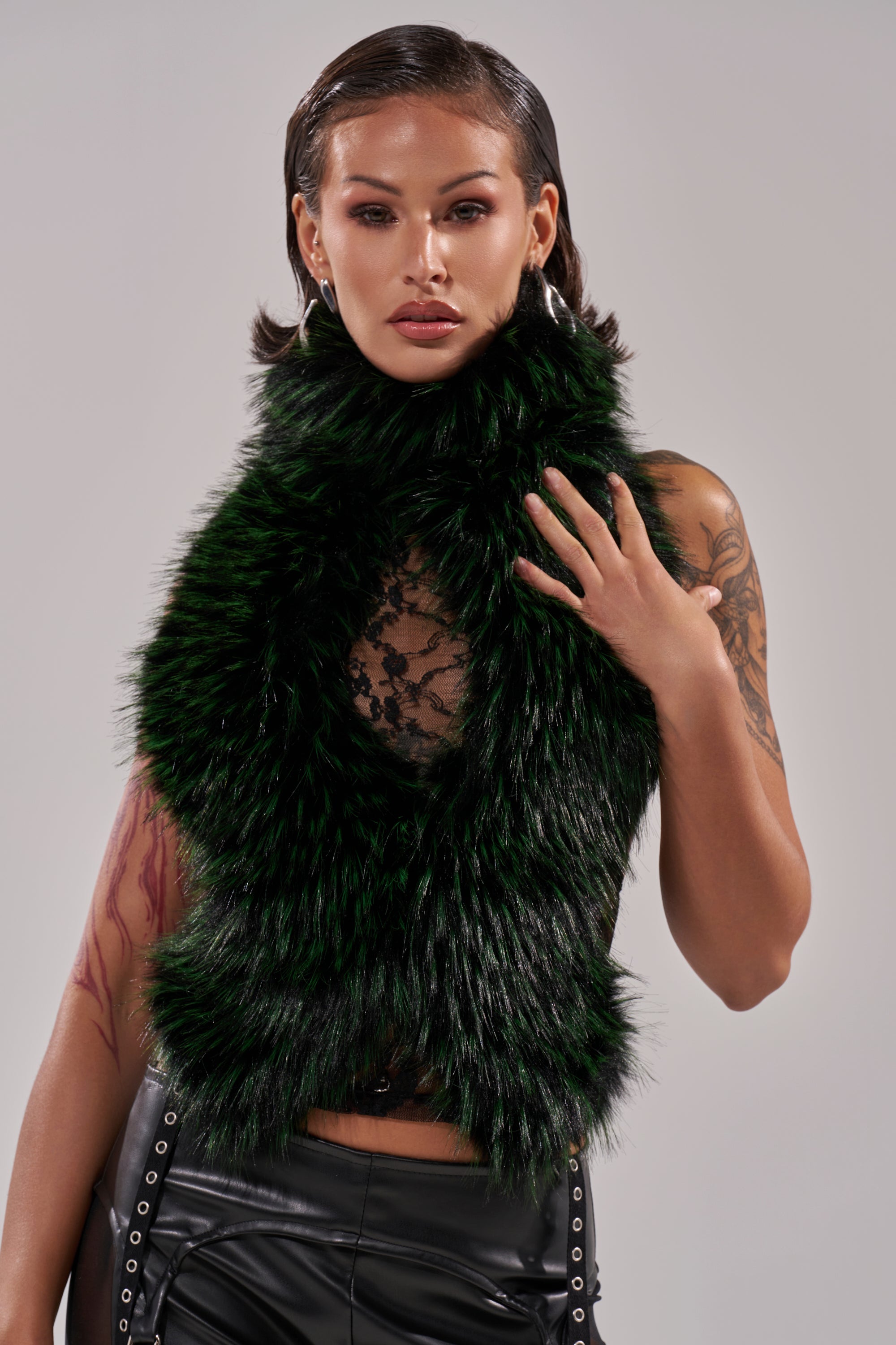 A person with slicked-back hair wears the FEEL ME BABY FUR BLOUSE in dark green, paired with black leather eyelet pants, hoop earrings, and sports arm tattoos. Their sheer lace top adds edge to the neutral background.