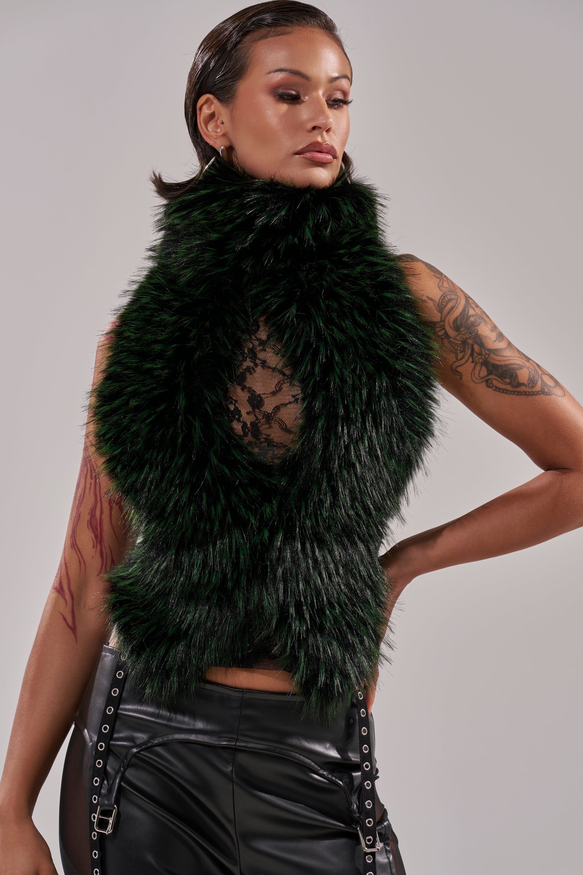 A woman with tattoos wears a sleeveless black lace top layered under the FEEL ME BABY FUR BLOUSE in dark green, paired with black leather pants featuring side grommet details. Her hair is slicked back.