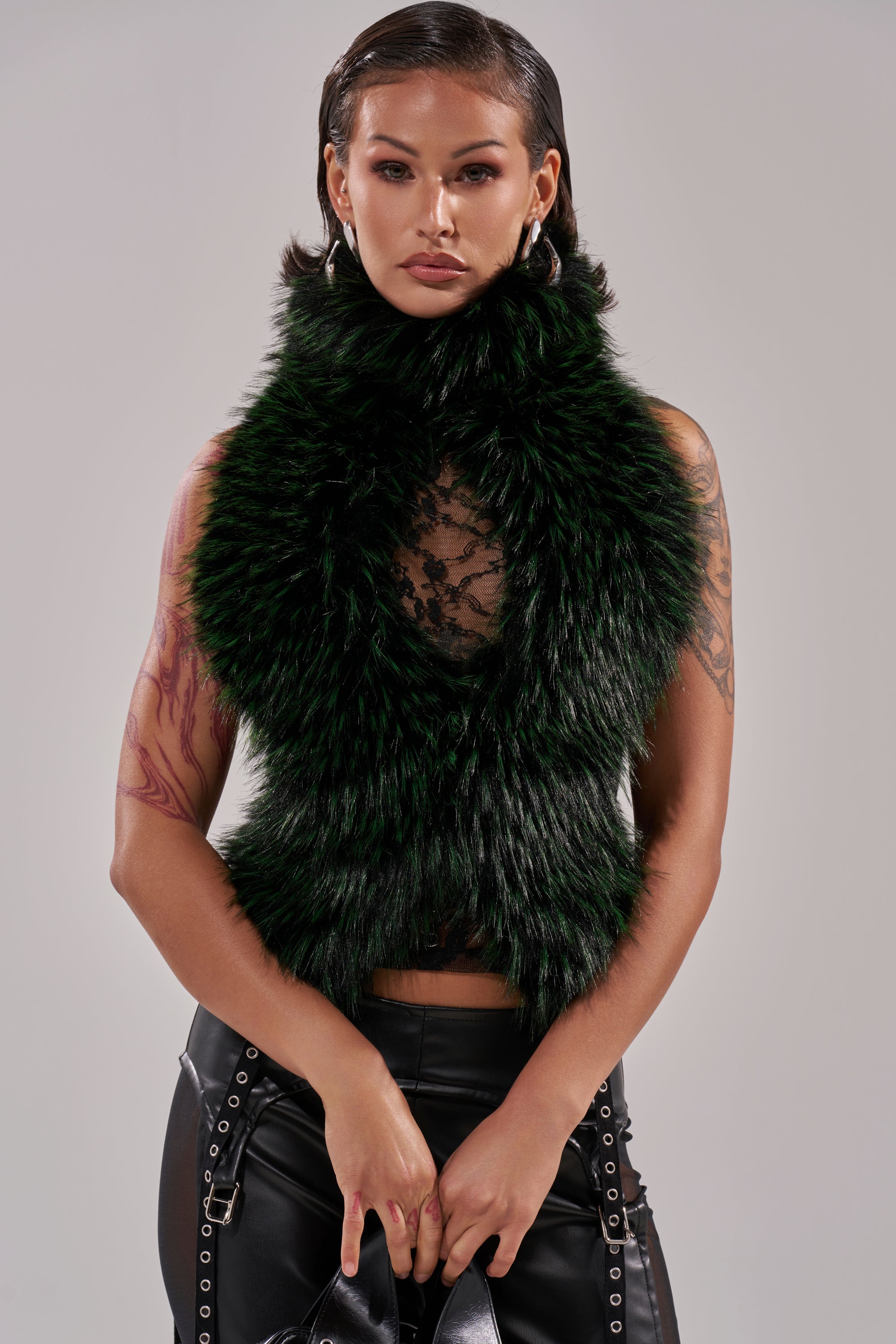 A woman with slicked-back hair wears the FEEL ME BABY FUR BLOUSE in dark green over a black lace top, paired with hoop earrings, black leather pants with grommet straps, visible arm tattoos, and holding a black bag.