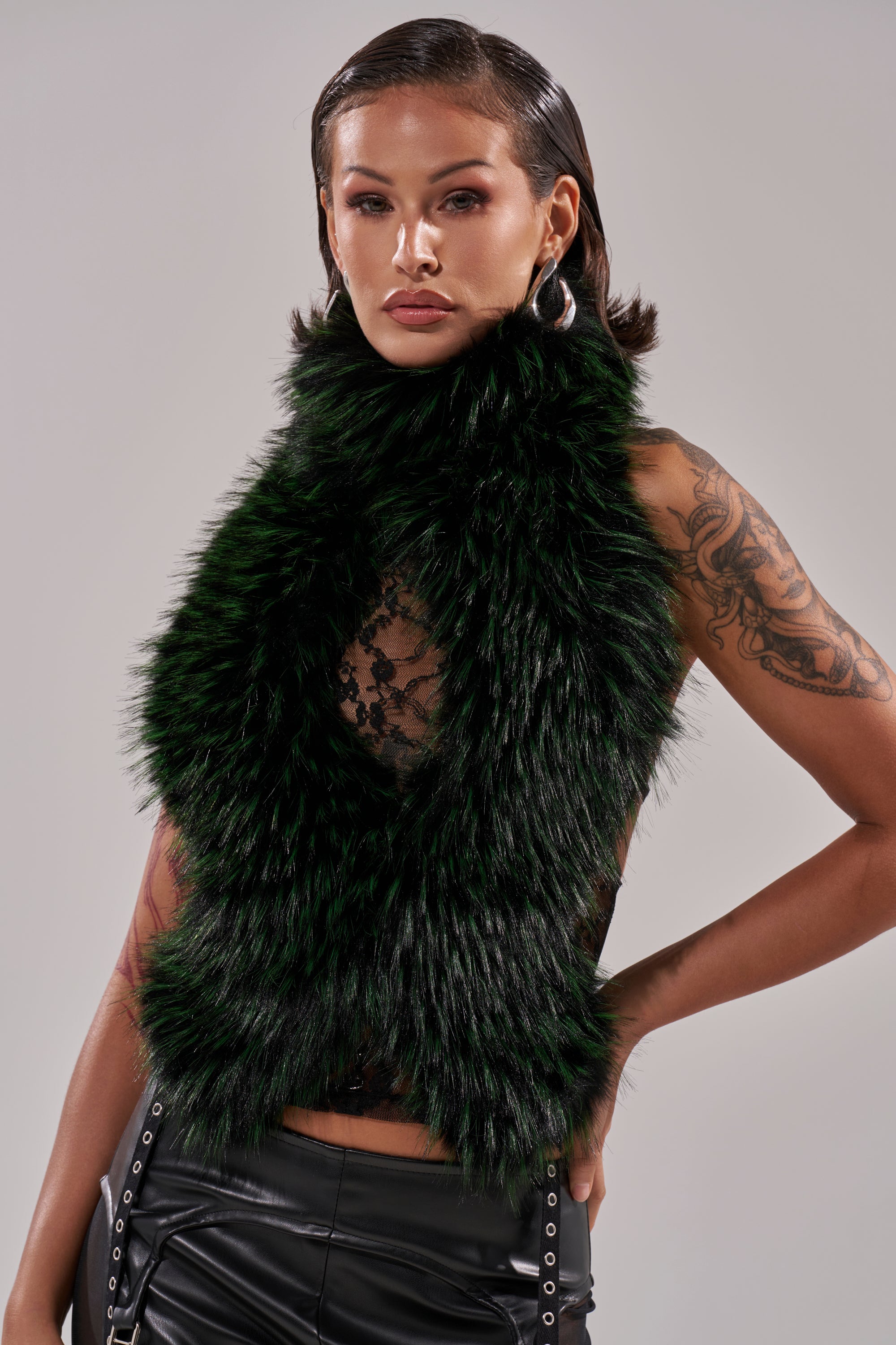 A woman with slicked-back hair and hoop earrings poses confidently in the FEEL ME BABY FUR BLOUSE—cropped, dark green—styled with a black sheer lace top, black leather pants, and showing arm tattoos against a light background.