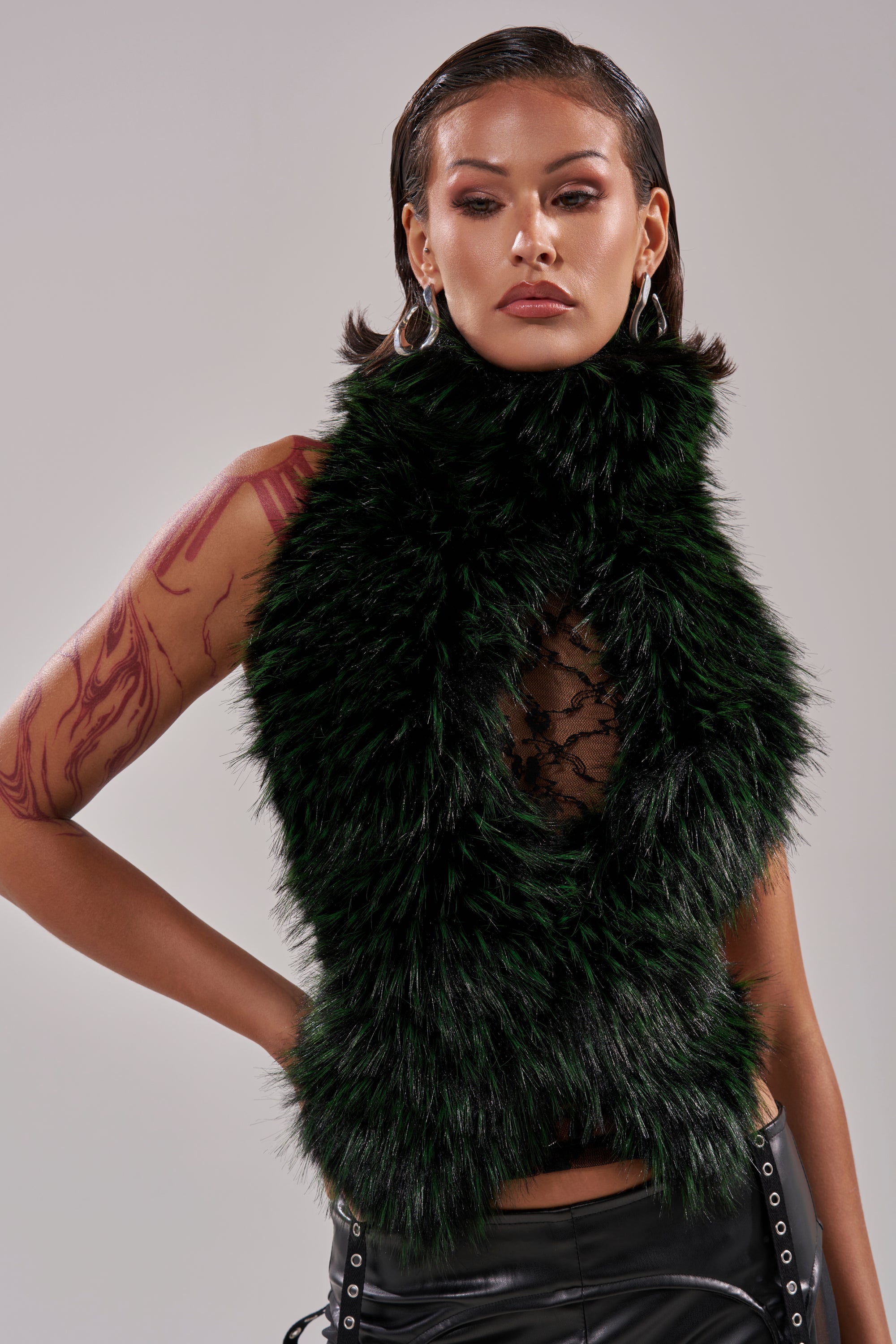A woman with slicked-back dark hair wears the FEEL ME BABY FUR BLOUSE—sleeveless, black, and faux fur—over a lacy top with hoop earrings and black pants. She poses confidently, showing bold makeup and a red tattoo on her right arm.