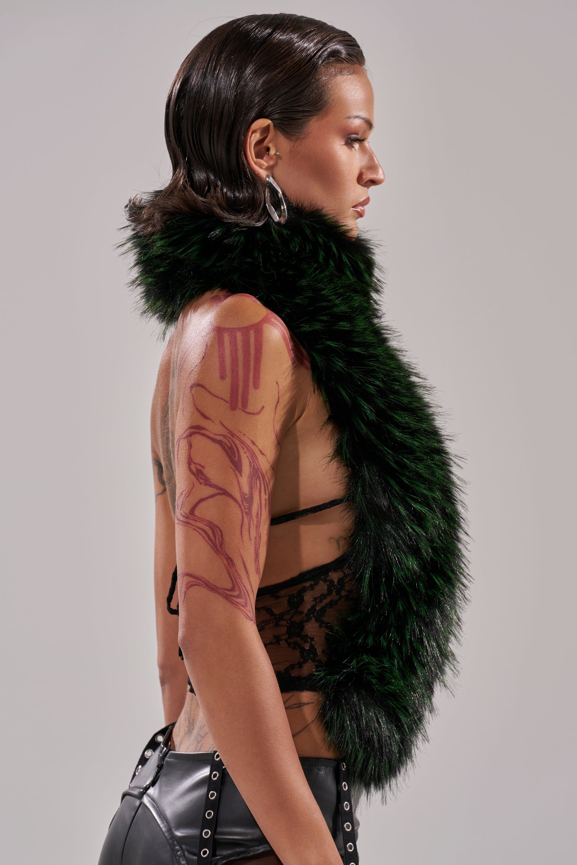 A person with slicked-back dark hair and abstract red tattoos wears the FEEL ME BABY FUR BLOUSE—a large, fluffy green ombré cropped top—with a black lace top and leather pants, standing in profile against a gray background.